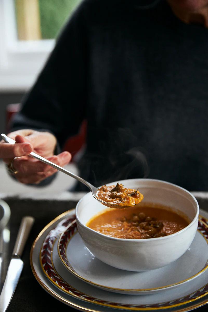 Someone in a dark sweater holds a spoonful of soup over a white bowl. The bowl sits on ornate plates in a naturally lit dining area.