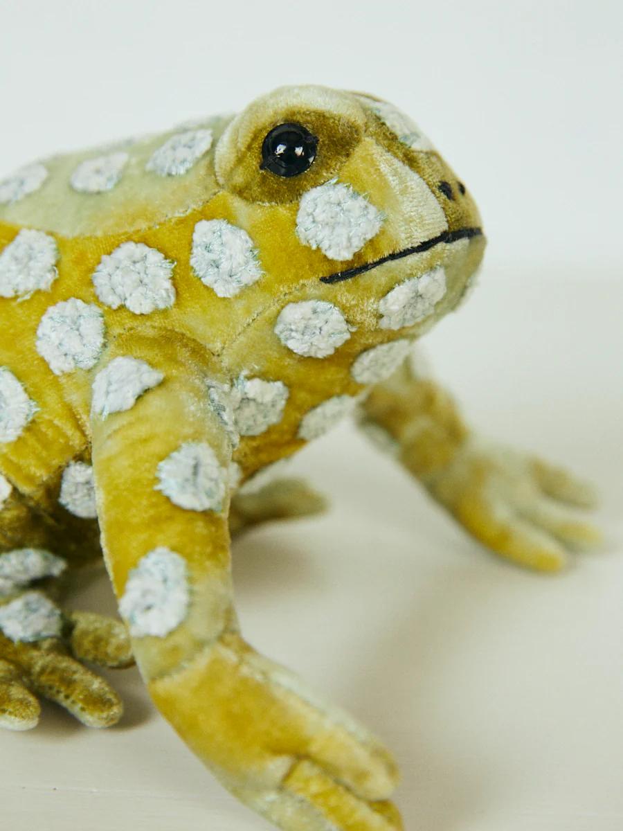 A plush toy toad with yellow-green skin and white spots sits on a light background. The realistic design features dark eyes and exquisite detailing.