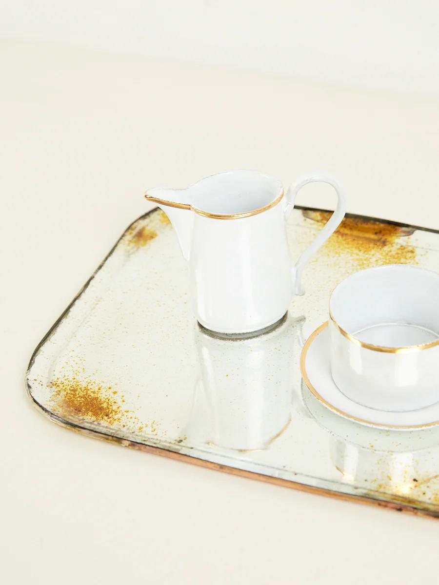 White ceramic milk jug, cup, and saucer on aged tray with tarnished spots. Soft beige background.