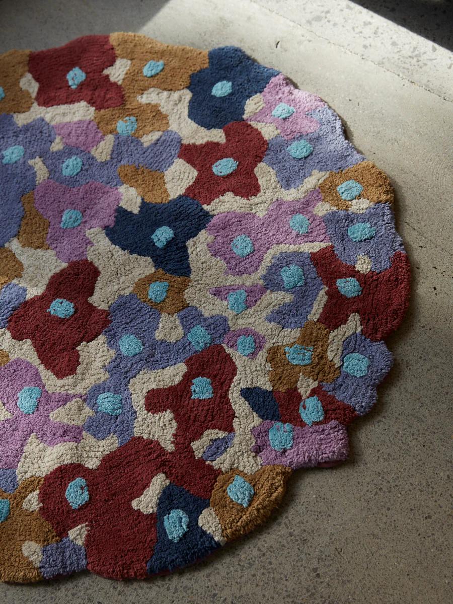 A round rug with an abstract floral pattern in red, blue, purple, and beige. Sunlight highlights the luxurious velour texture on the concrete floor.