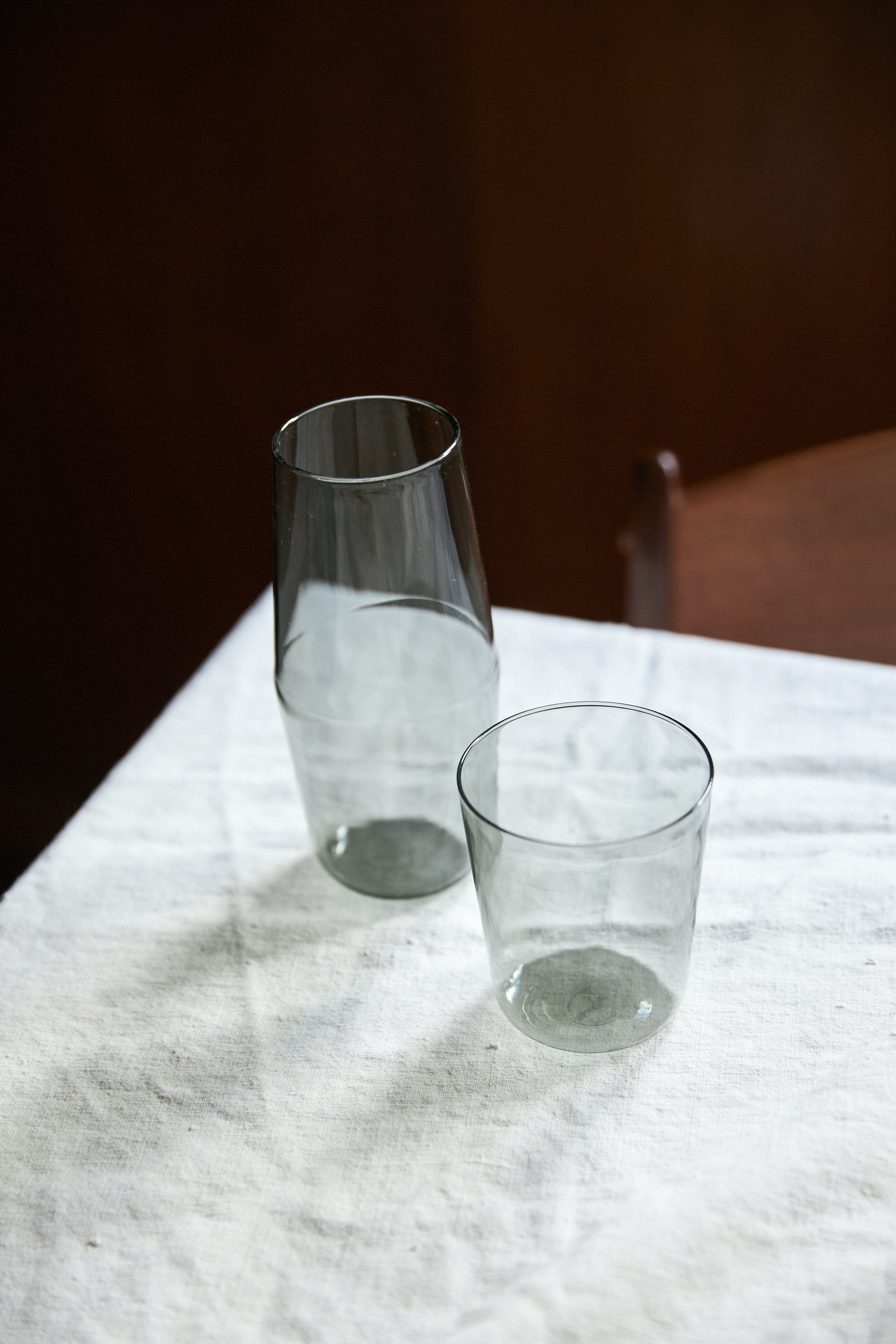 A grey-tinted carafe and glass set on a white tablecloth. A dark wooden chair is visible in the background.