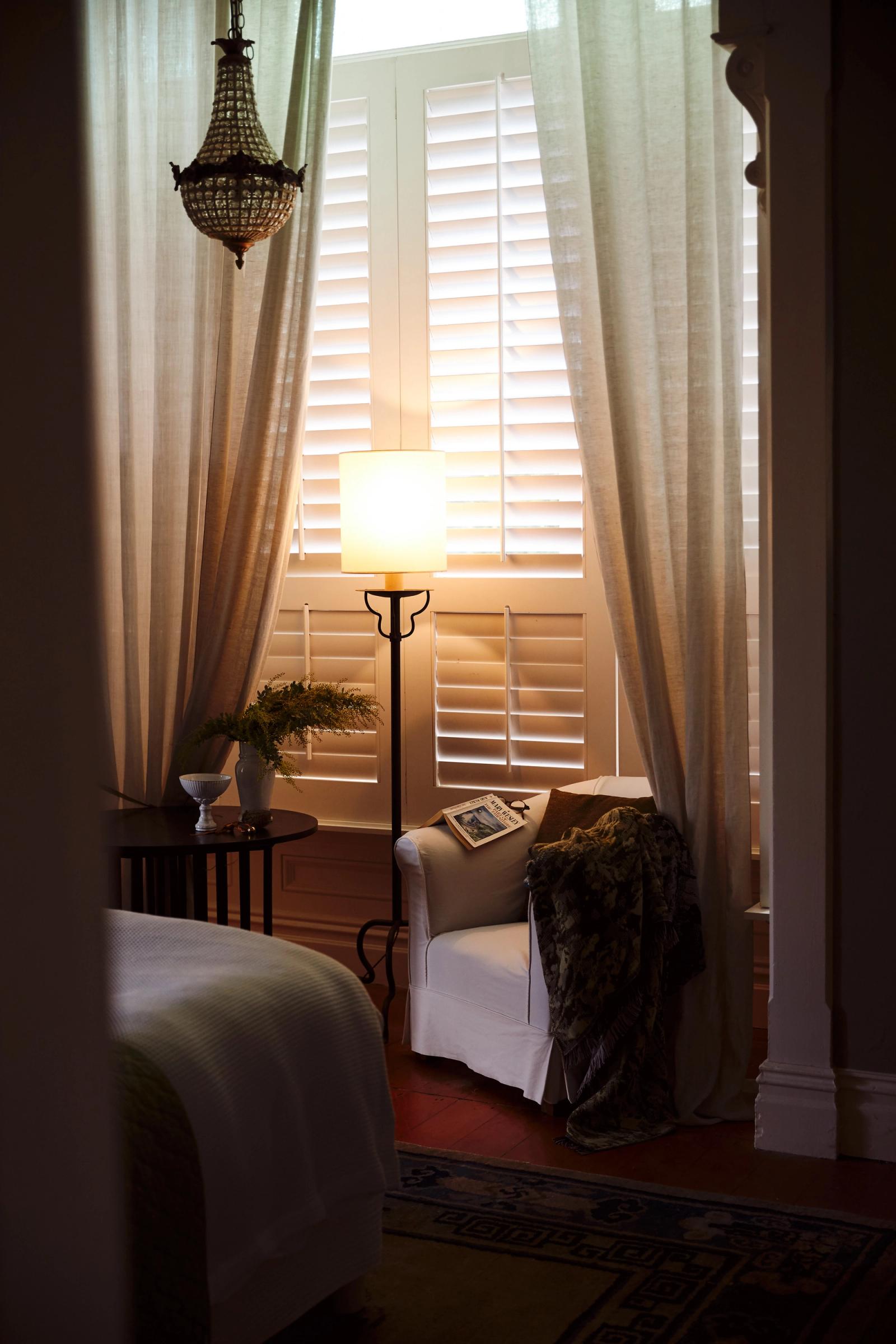 lamp glows next to shutter and sheer curtains in a bedroom 