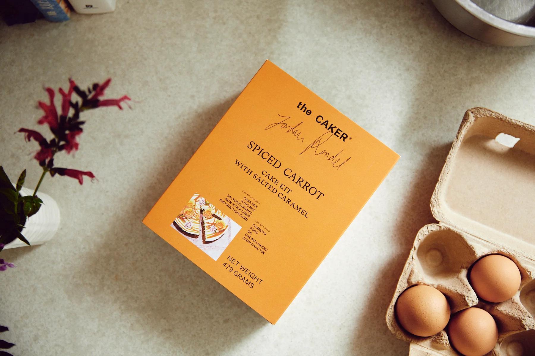 Orange "Spiced Carrot Cake Kit" box near egg carton. Partial flower bouquet and metallic bowl visible.