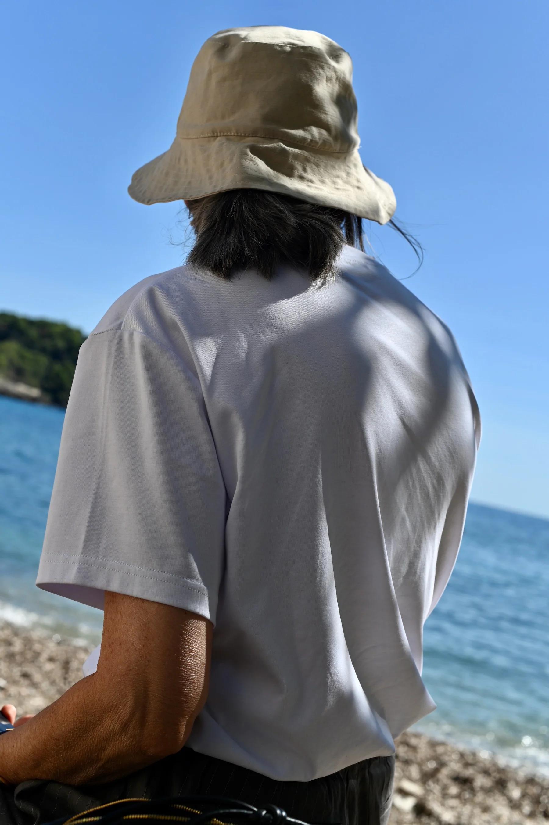 Person in white T-shirt and beige hat facing sea. Clear blue sky, calm water, and pebbled shoreline. Trees visible in distance.