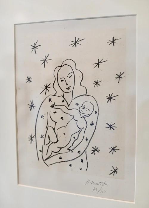 A black and white drawing depicts a woman holding a baby surrounded by stars. The minimalist style uses simple lines to form the figures. This framed artwork is signed "H. Matisse 76/100,".