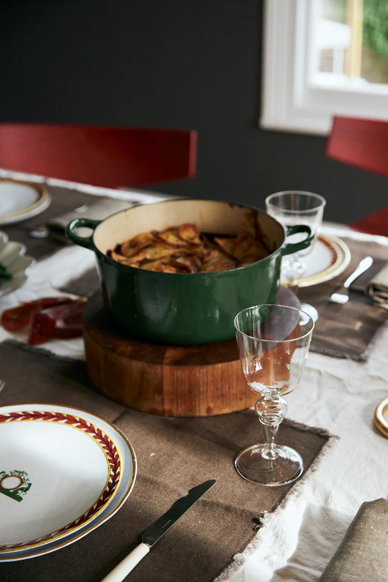 Green pot with beef casserole on wooden board. Table set with plates, wine glasses, napkins. Red chairs in background.