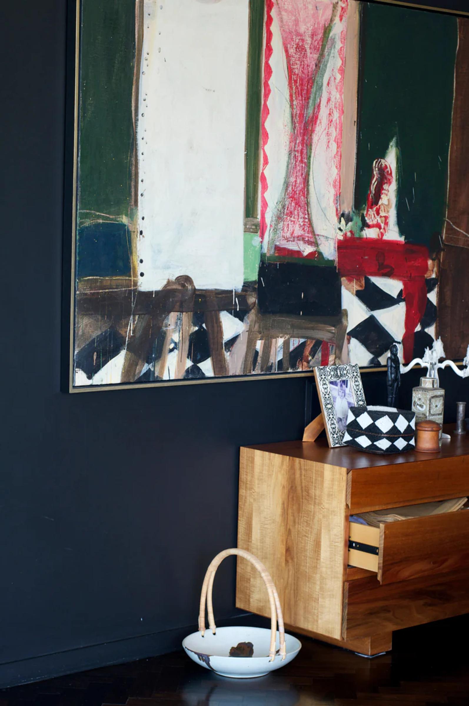 Abstract painting with bold colours above wooden sideboard. Decorative items, framed photos, and checkered box on sideboard. Basket on floor nearby.