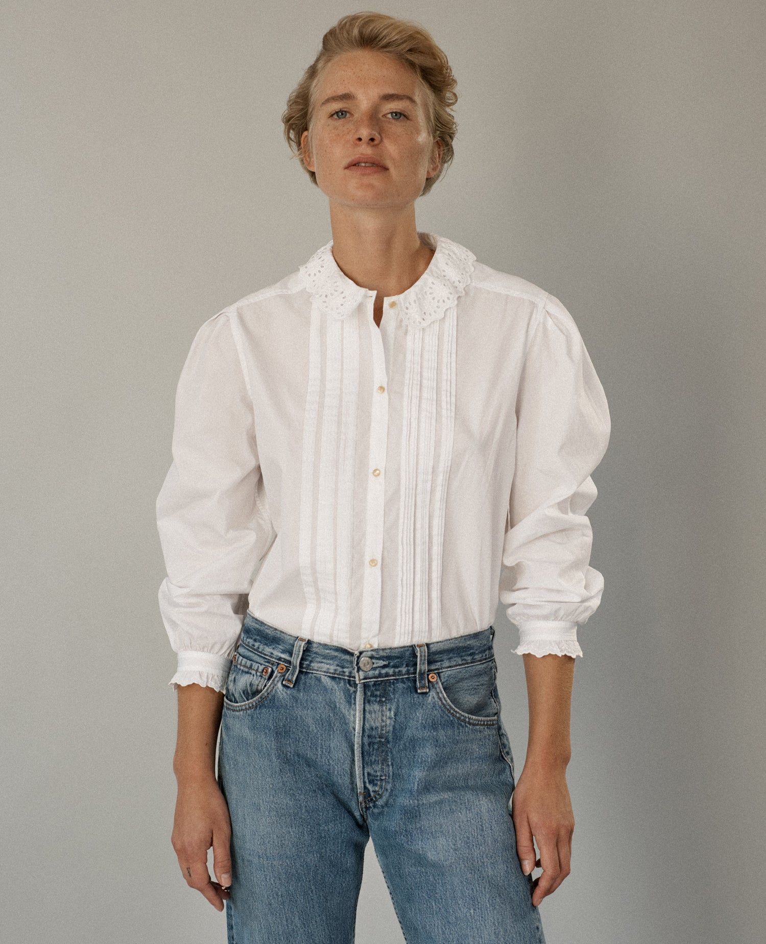 Person in white long-sleeved blouse with decorative collar and jeans. Standing against grey background, looking upward.