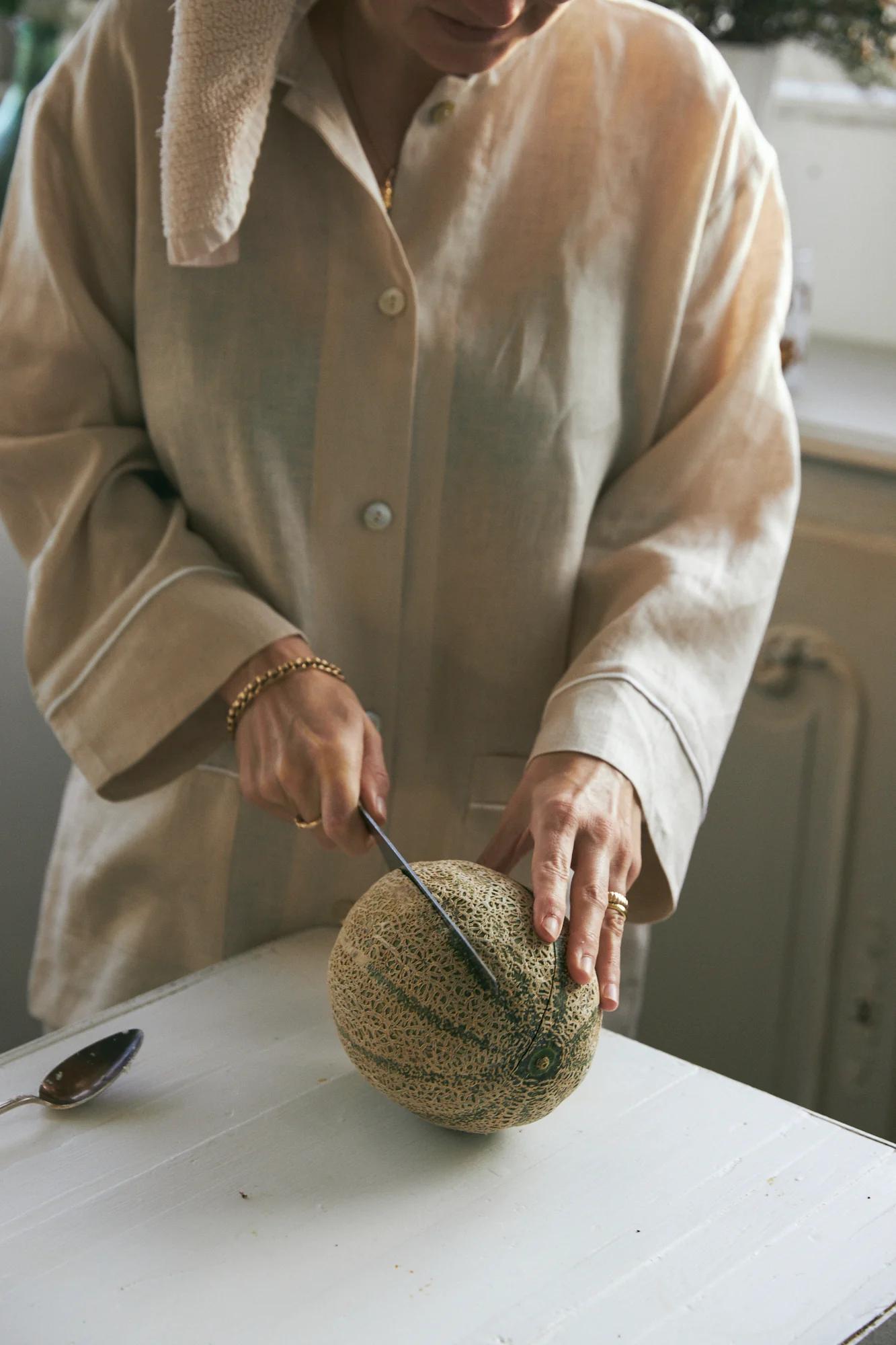 Person in beige shirt slicing cantaloupe on white table. Knife, spoon, and shoulder-draped towel visible. Captures lazy breakfast vibe.