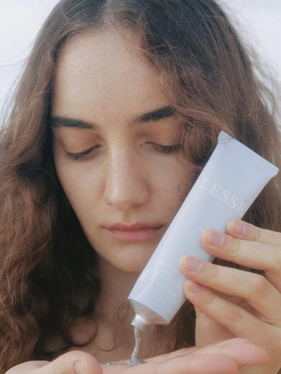 Person with wavy hair squeezes LESSE cream onto hand. Soft, light background emphasises organic skincare focus.