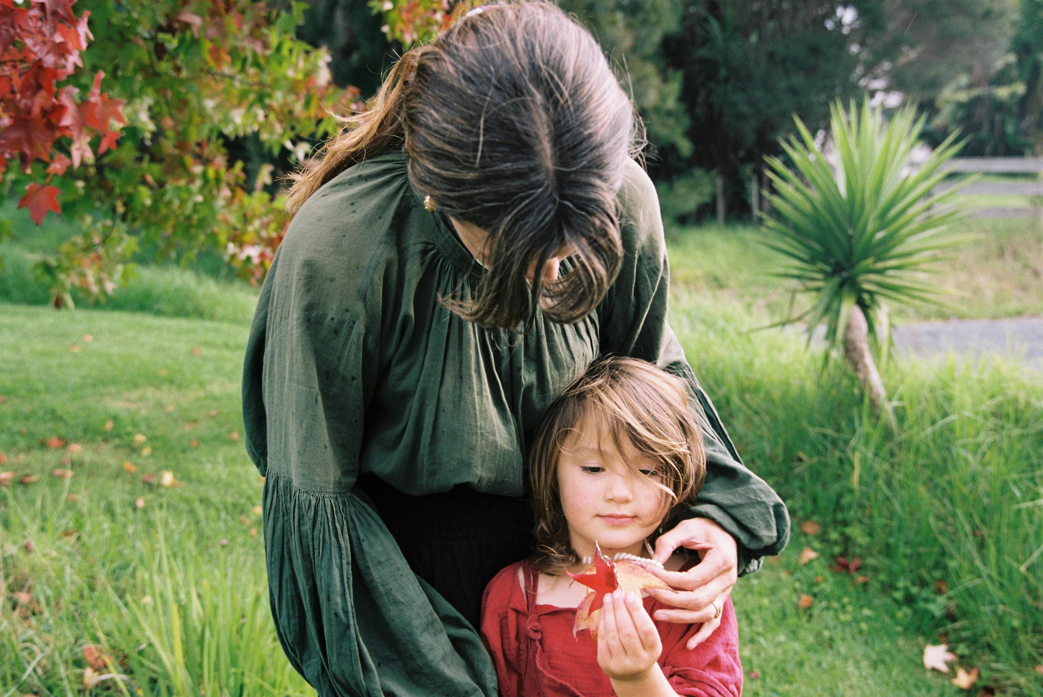 A woman in a green dress bends over to embrace a child holding a red leaf. The child in a red dress stands on grass with trees behind them, capturing a sweet moment.