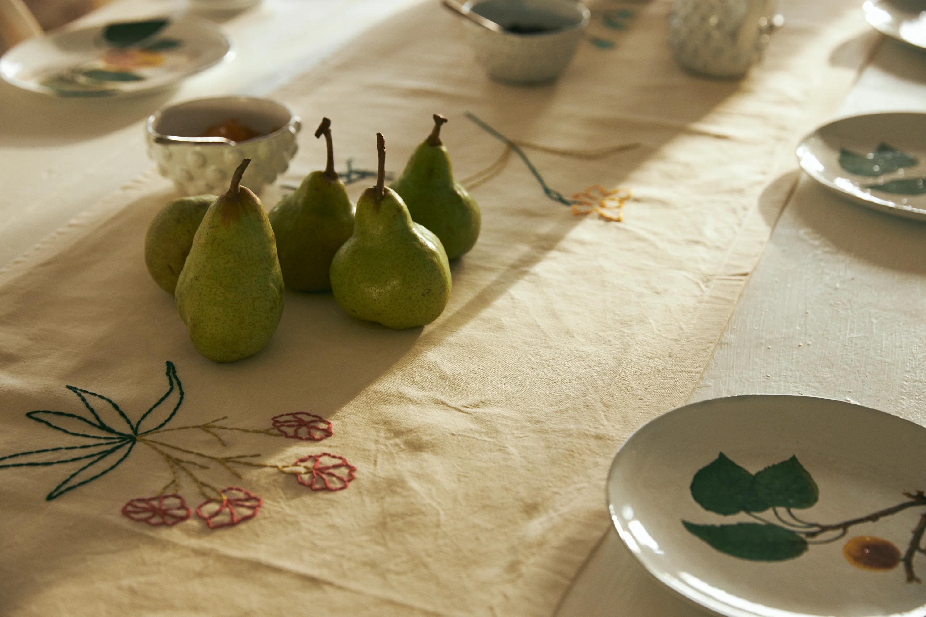 Table with embroidered runner and green pear centrepiece. White plates and bowls with leaf designs. Perfect for cosy holiday breakfast.