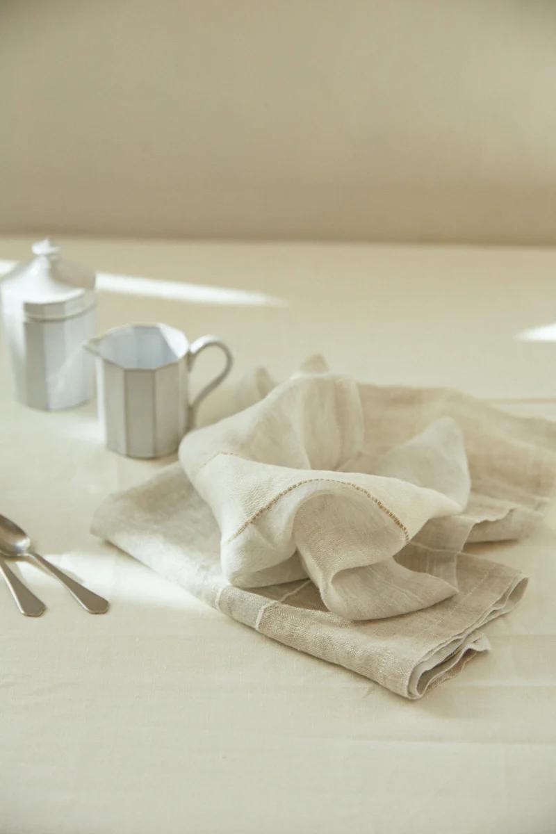 Light beige Libeco napkins, metal milk jug, sugar container, and teaspoons on light tablecloth. Soft sunlight ambiance.