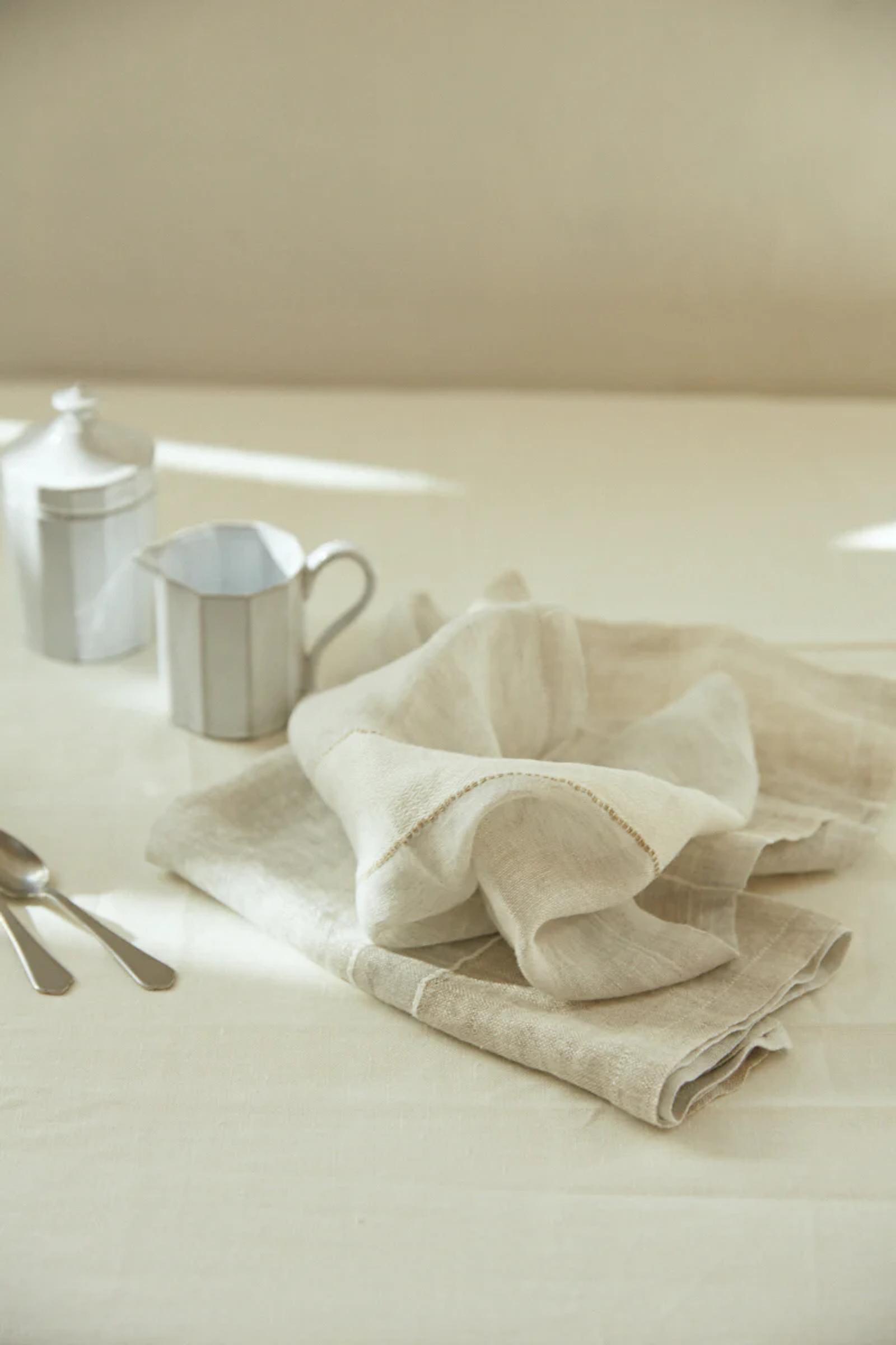 Light beige Libeco napkins, metal milk jug, sugar container, and teaspoons on light tablecloth. Soft sunlight ambiance.