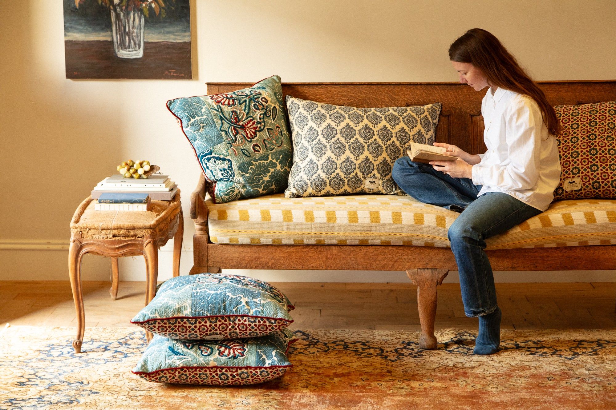 Woman reading on cushioned bench. Table with books nearby. Large cushions on floor. Framed painting on wall behind.