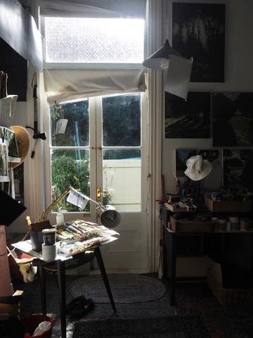 Cosy Devonport studio with sunlit glass door. Desk laden with paints and brushes. Walls adorned with Thomson's paintings and sketches. Hat hangs by door.