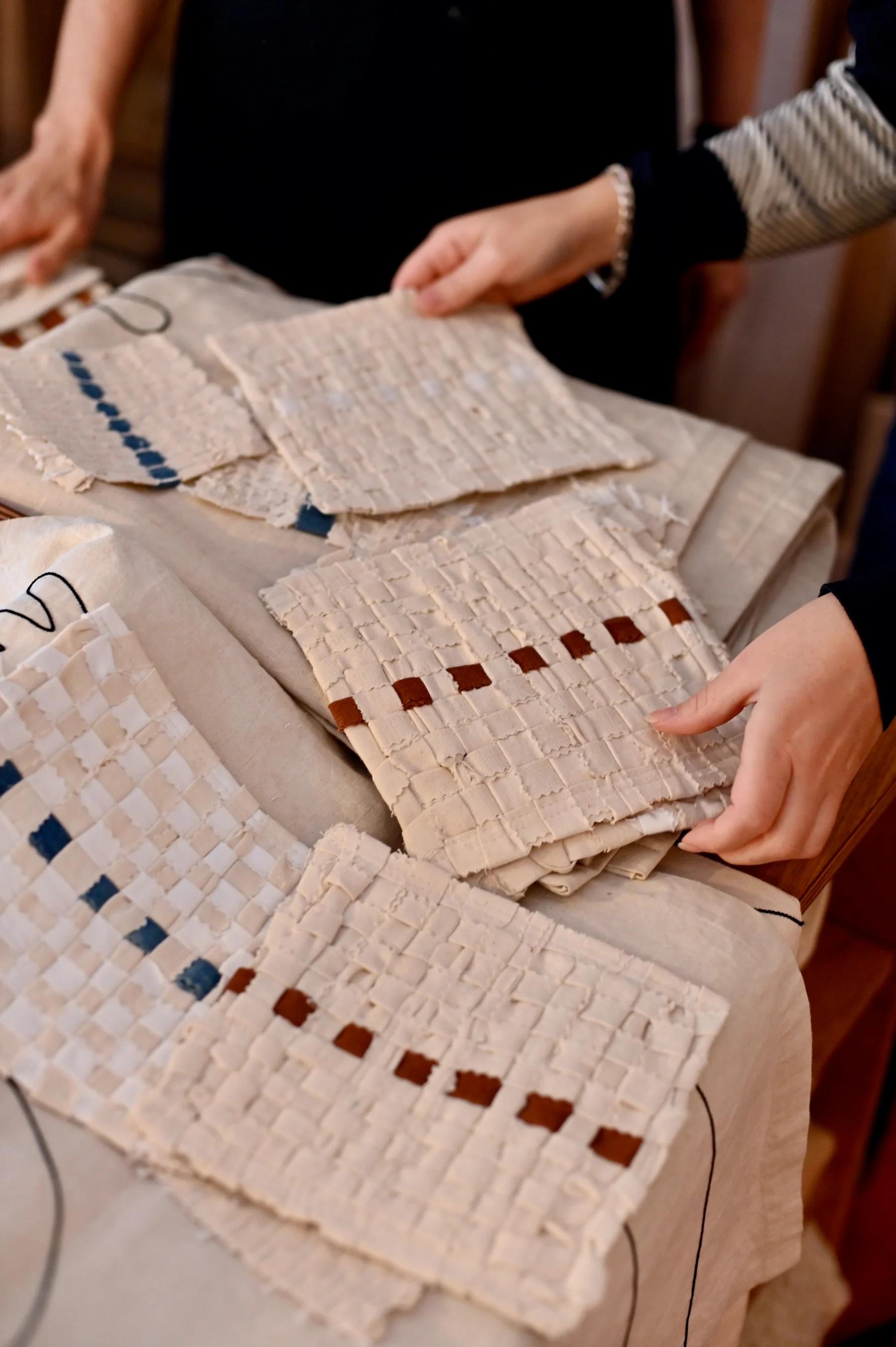 Two people examine textiles on table. Fabrics show intricate weave patterns in neutral tones with blue and brown accents.