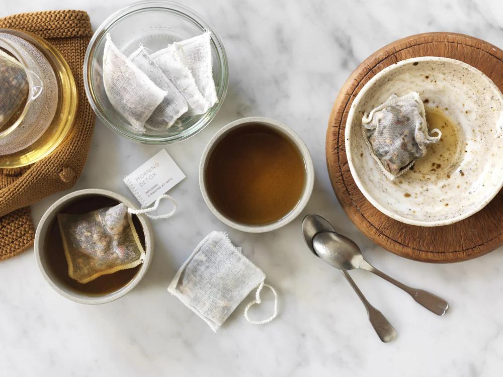 Marble surface with white ceramic containers holding teabags. One cup of tea, vintage teaspoons, saucer, and glass bowl with multiple teabags.