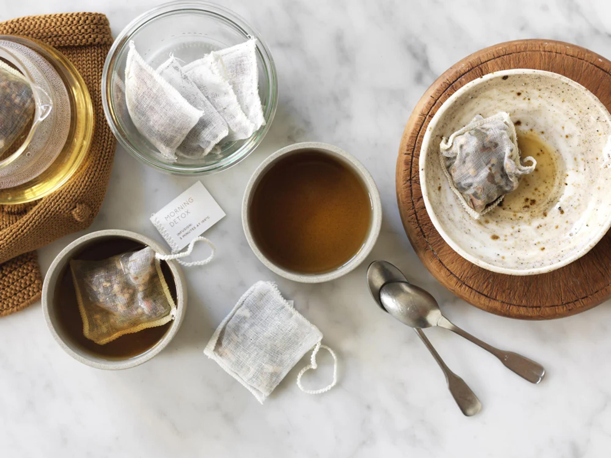 Marble surface with white ceramic containers holding teabags. One cup of tea, vintage teaspoons, saucer, and glass bowl with multiple teabags.