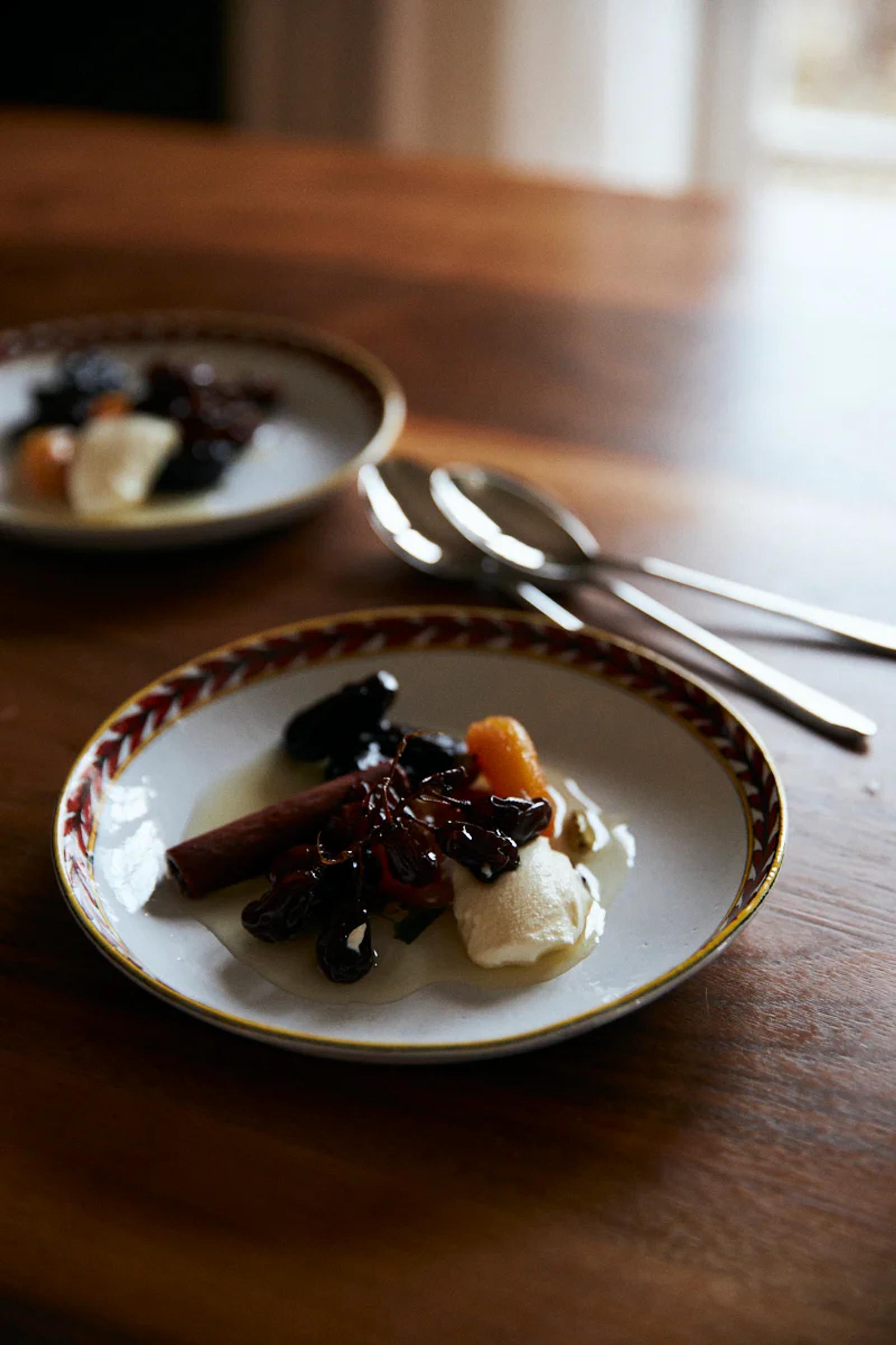 White plate with red-and-gold rim holding fruits and cream on wooden table. Two spoons nearby, similar plate blurred in background.