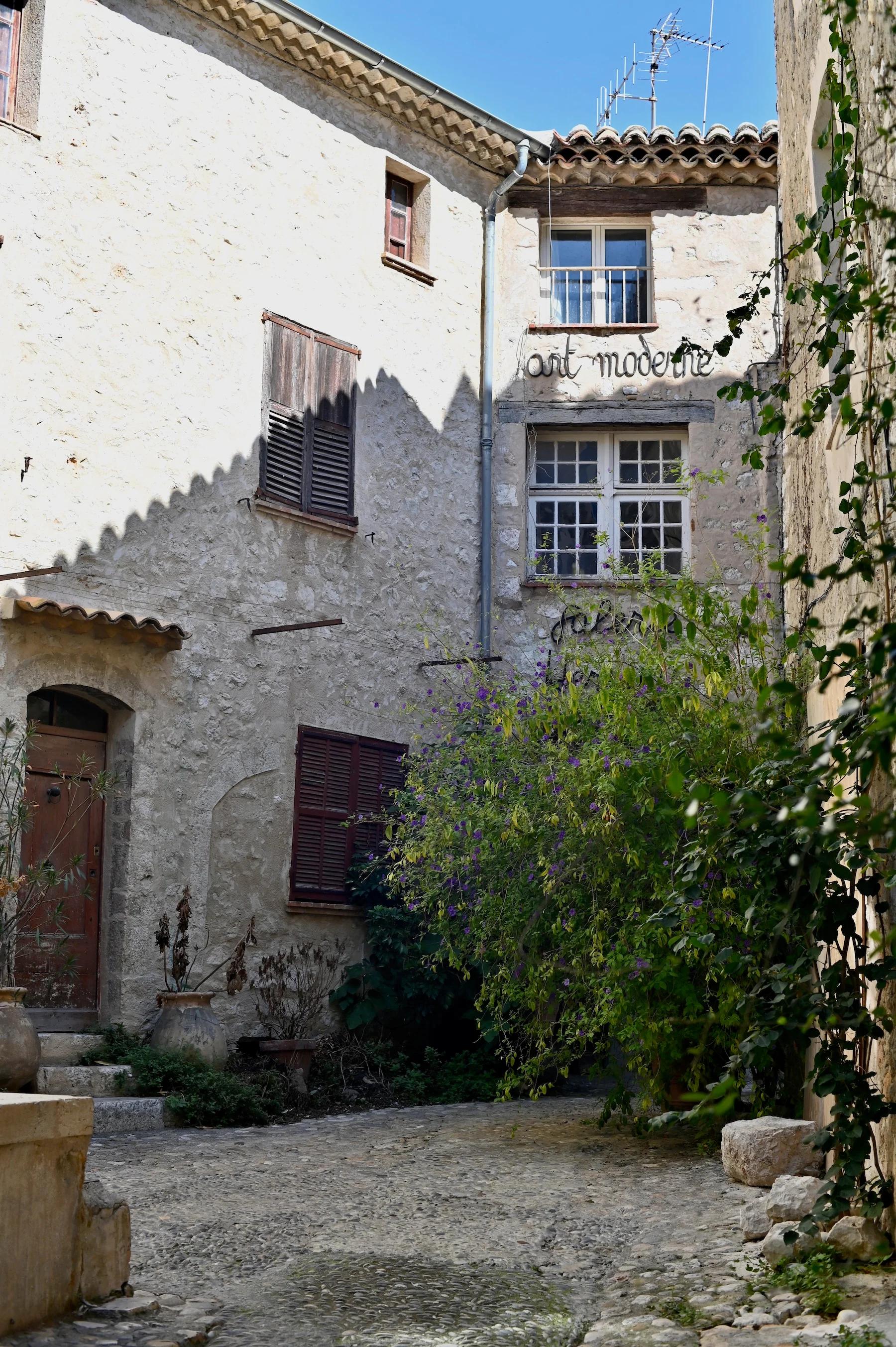 Narrow sunlit courtyard with stone buildings and "art moderne" signs. Purple flowers grow against a wall, with cobblestone paths evoking South of France charm.