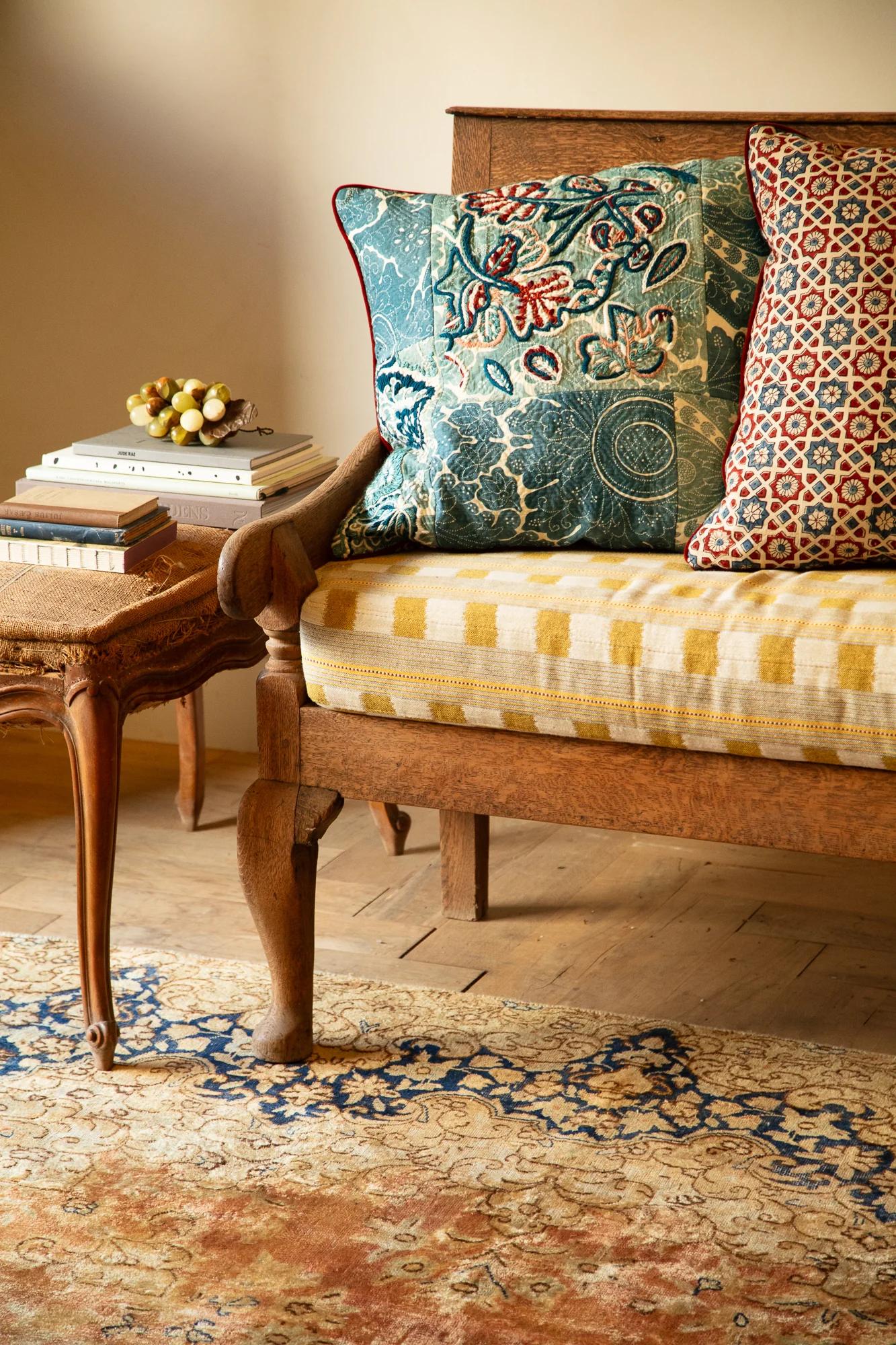 Rustic living room. Wooden bench with yellow cushions, patterned pillows. Side table with books, grapes. Patterned rug on floor.