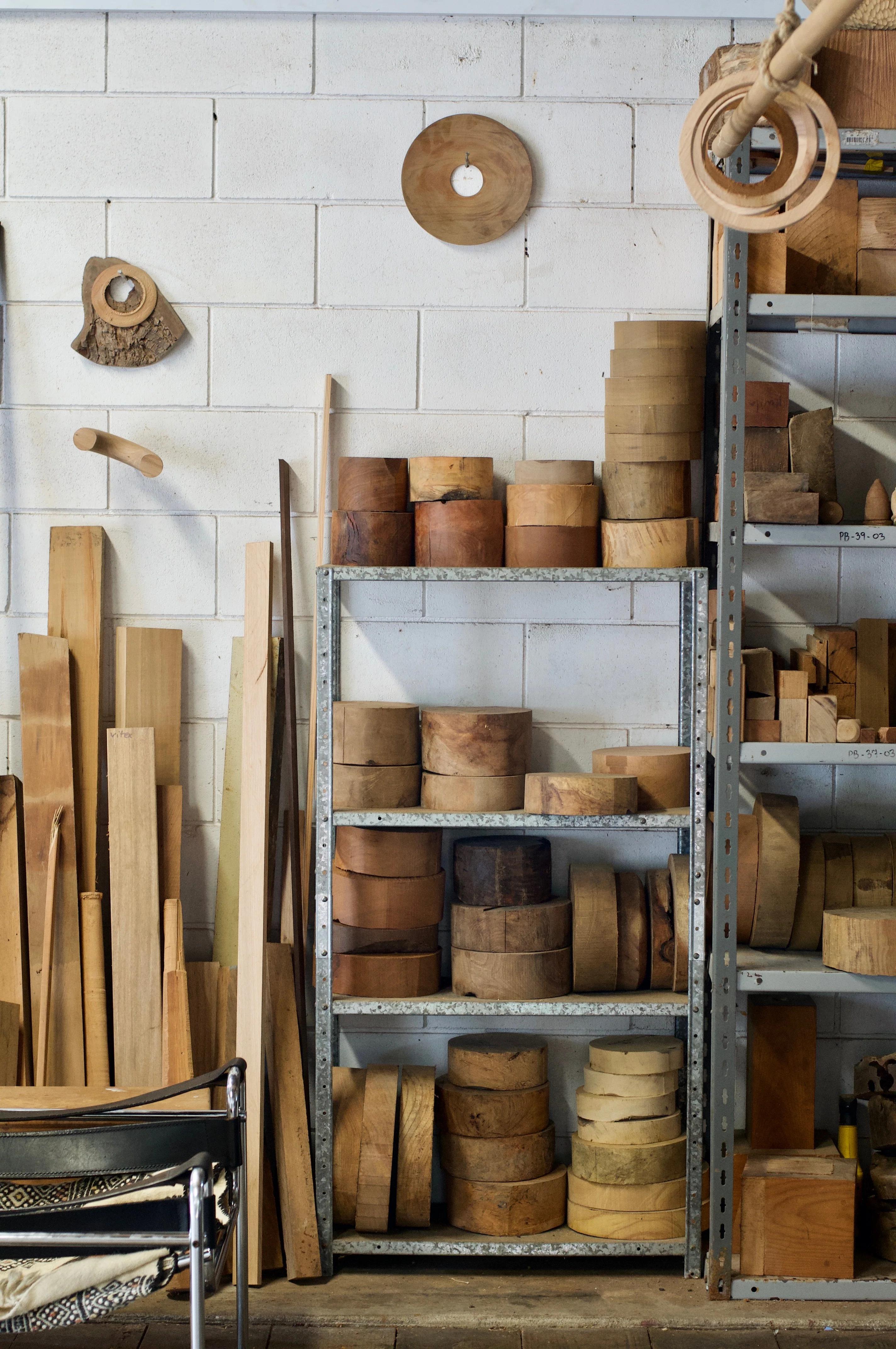 Workshop with stacked wooden logs and planks on metal shelves. White brick wall backdrop. Various woodworking materials and tools, including circular pieces and molds, from NZ wood on floor and shelves.