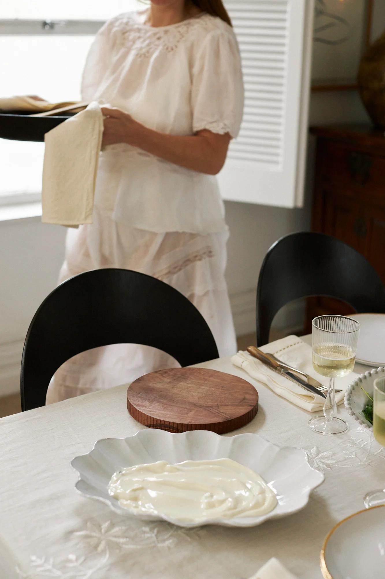 A dining table set for Easter with a wooden board, ornate dish, utensils, and wine. A person in white holds a tray in the background.