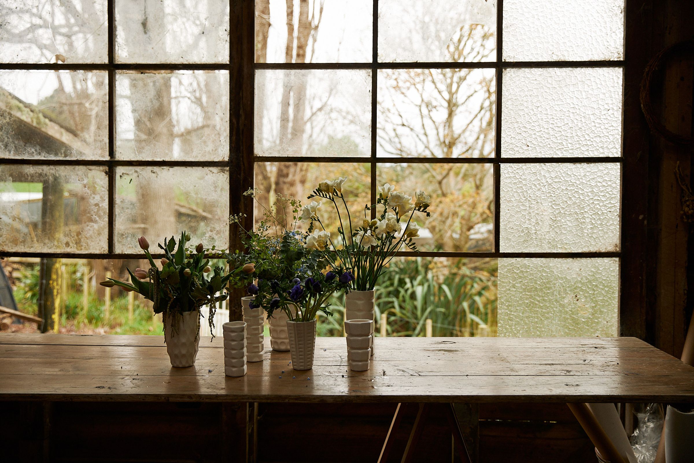 line up of vases with flowers sits on table against glass window panes