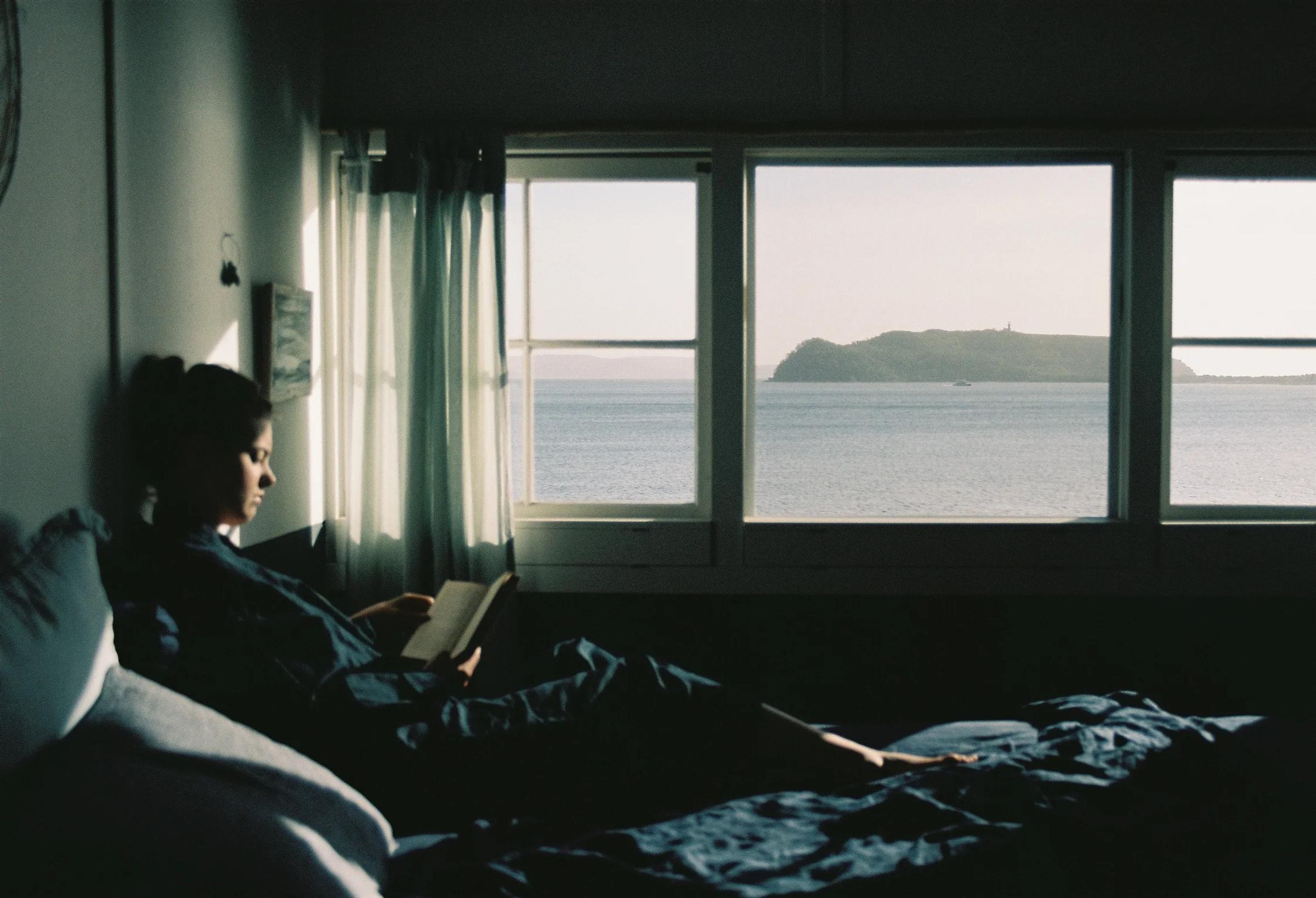 A person reads a Thich Nhat Hanh book in bed. Sunlight streams through a window framed with white curtains, revealing an ocean view and distant island, creating a peaceful scene.