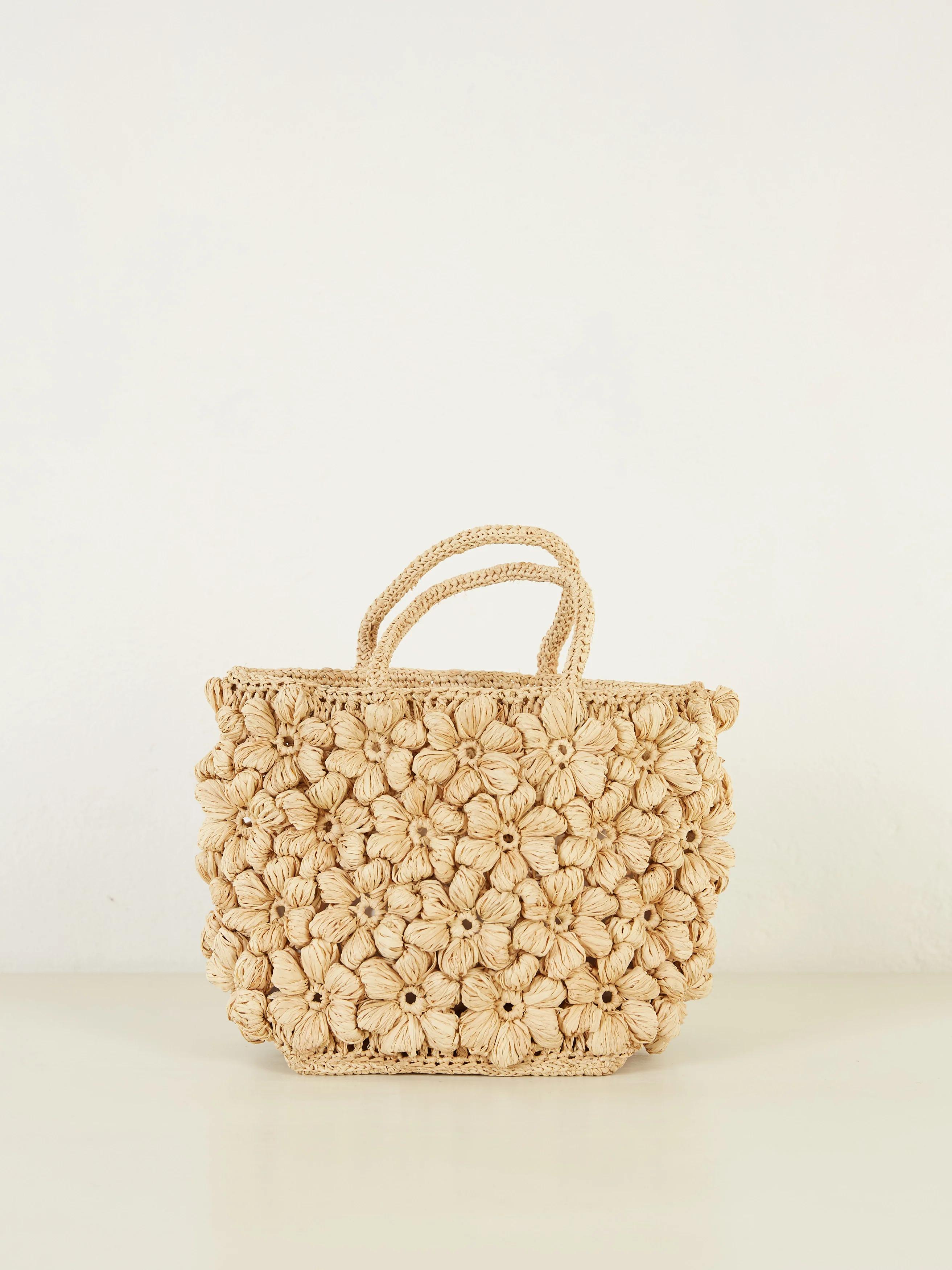 A beige woven raffia handbag with a flower pattern, matching handles, and a neutral background.