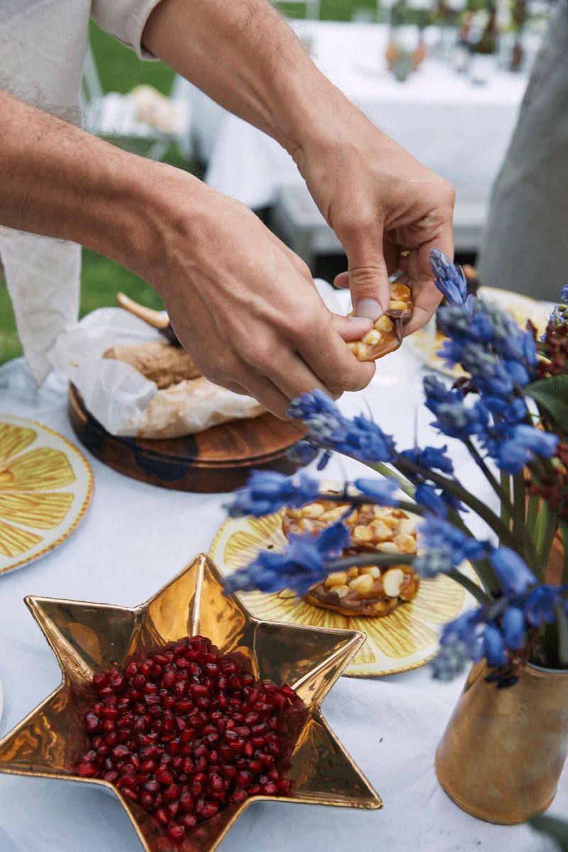 Person breaking candied brittle over table. Star-shaped bowl with seeds, vase of blue flowers, and table settings.