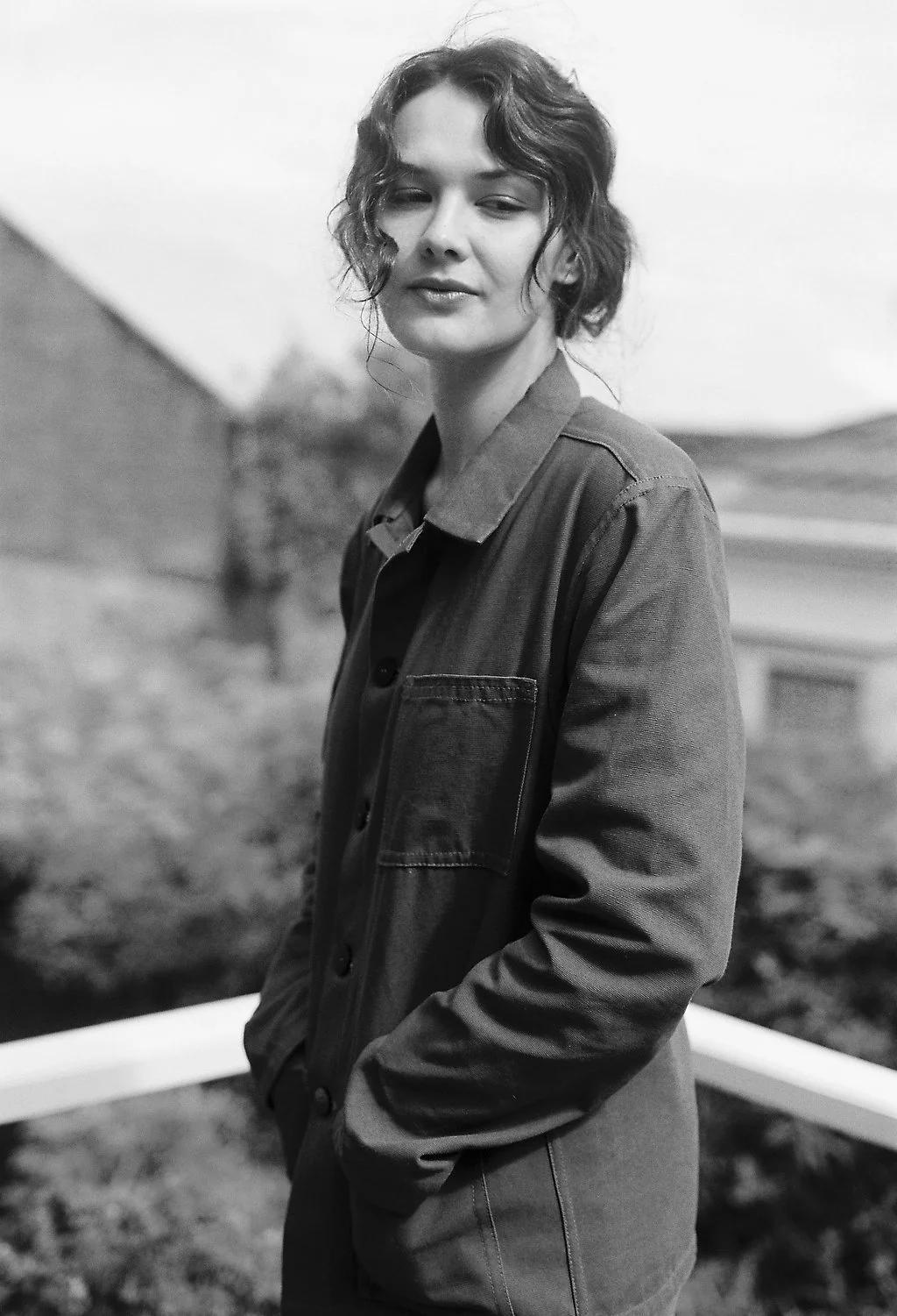 Black and white photo of curly-haired person in buttoned jacket. Standing outdoors, hands in pockets, gazing away. Blurred building and greenery behind.