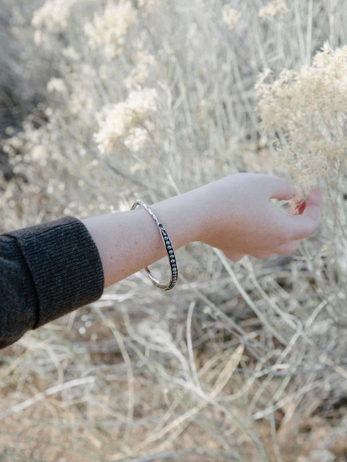 Outstretched arm wearing black sleeve and two bracelets against dry grass. Natural light highlights outdoor setting, reflecting organic-inspired jewellery design.