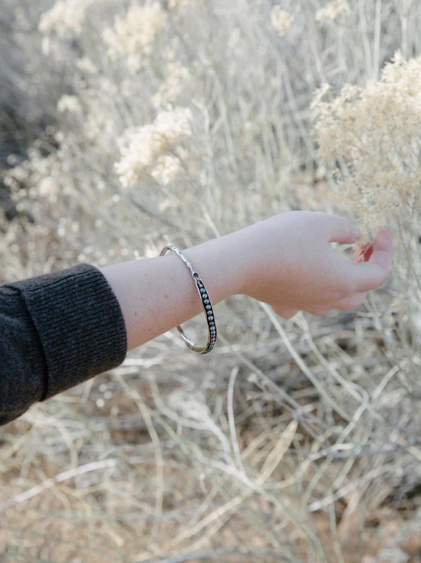 Outstretched arm wearing black sleeve and two bracelets against dry grass. Natural light highlights outdoor setting, reflecting organic-inspired jewellery design.