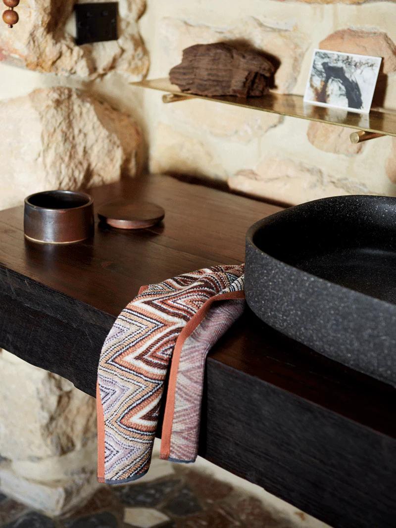 A stone basin on a wooden counter with a patterned towel draped over the edge, beside two round containers and a stone backsplash.