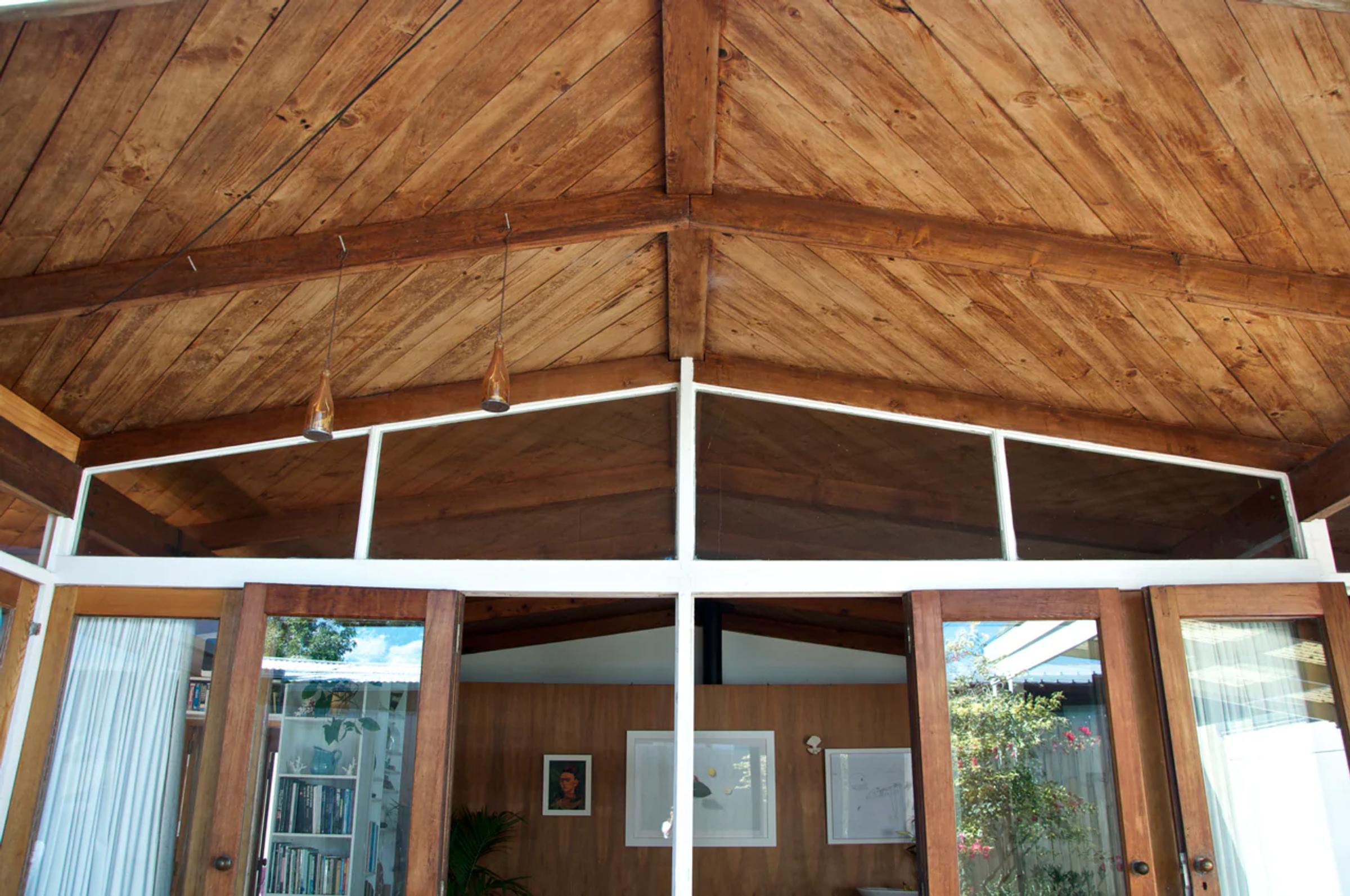 Living space with wooden pitched ceiling and beams. Large glass doors and windows. Bookshelf with collections and framed pictures on wall. Minimalist, cosy atmosphere.