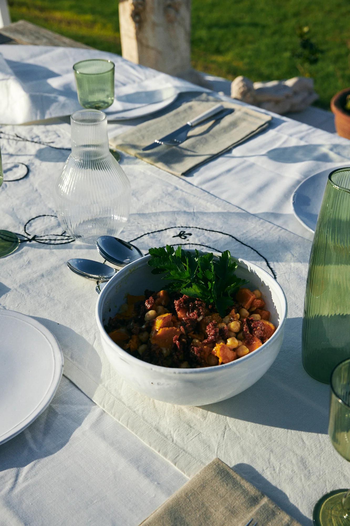 White bowl with colourful stew on outdoor table. Table set with plates, glasses, cutlery. Green lawn in background.