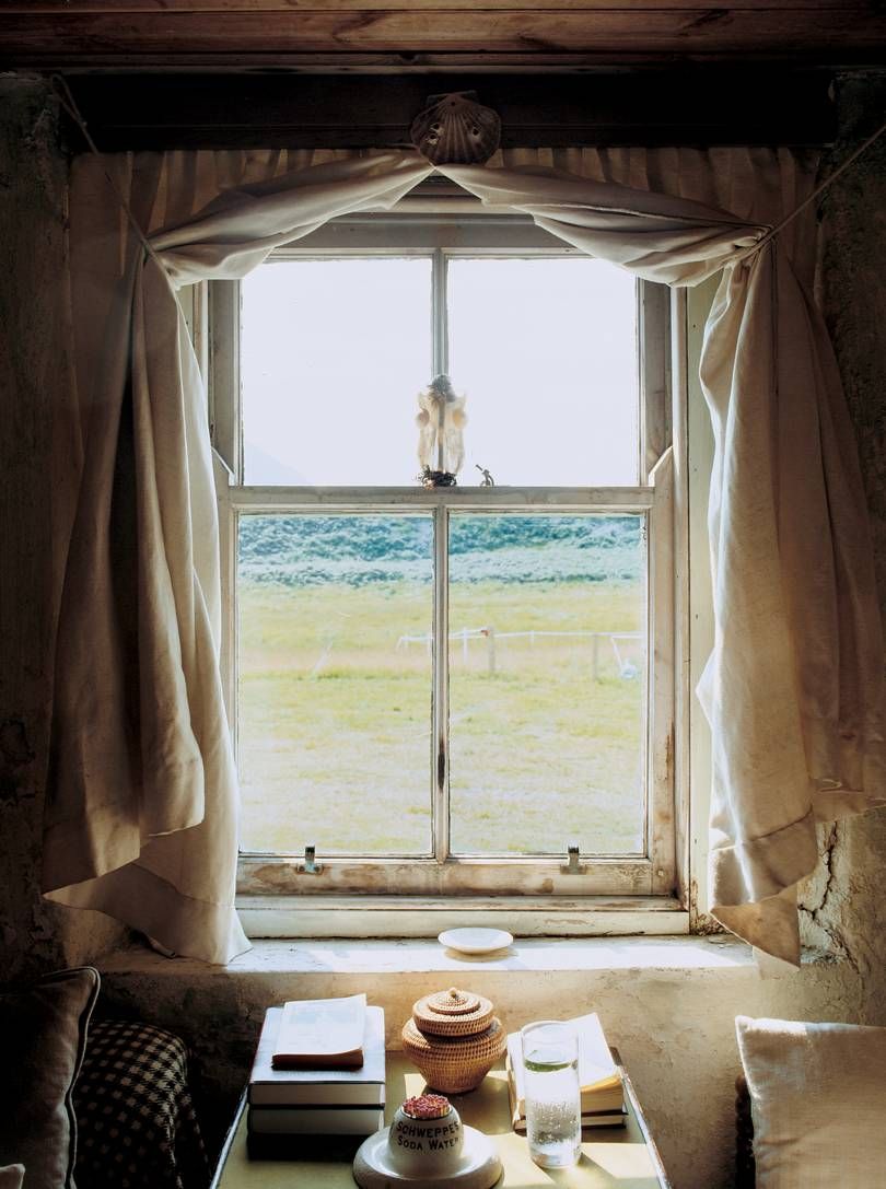 Rustic window overlooking green field. Table below holds wicker jar, books, and sugar jar. Sunlight streams in. Jura-like scene.