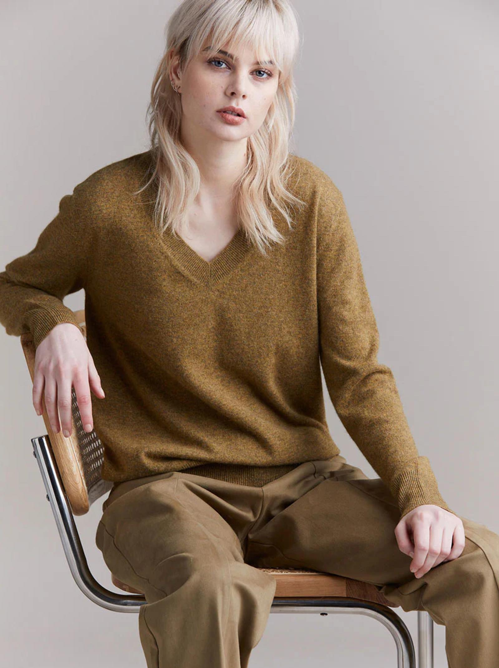 Blonde person in olive sweater and khaki pants on wooden chair. Relaxed pose against light grey backdrop.