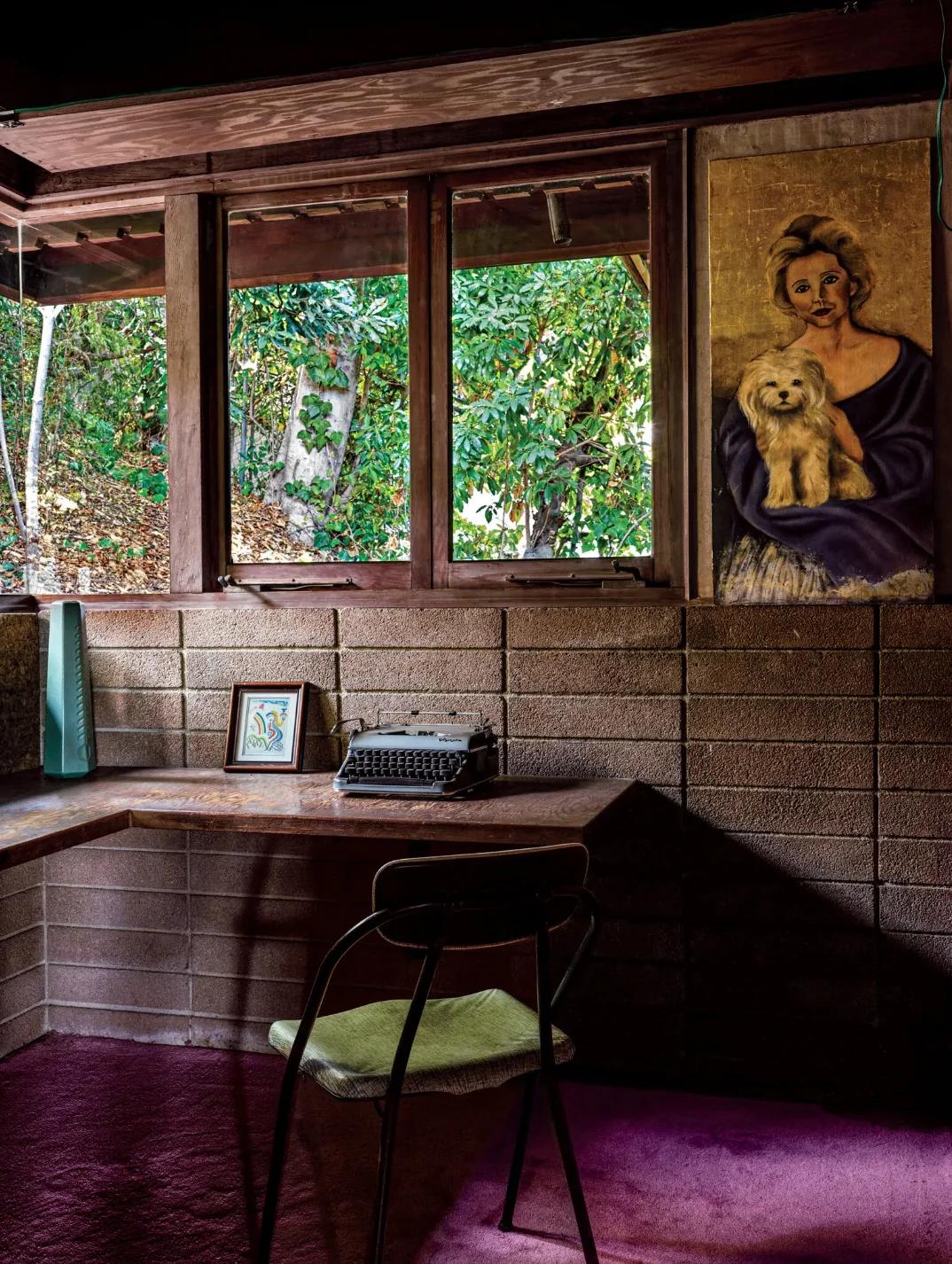 A cozy Los Angeles home workspace with desk and brick walls. Features a vintage typewriter, a framed picture, chair, and hanging portrait of a woman and dog. Greenery is visible through the window.