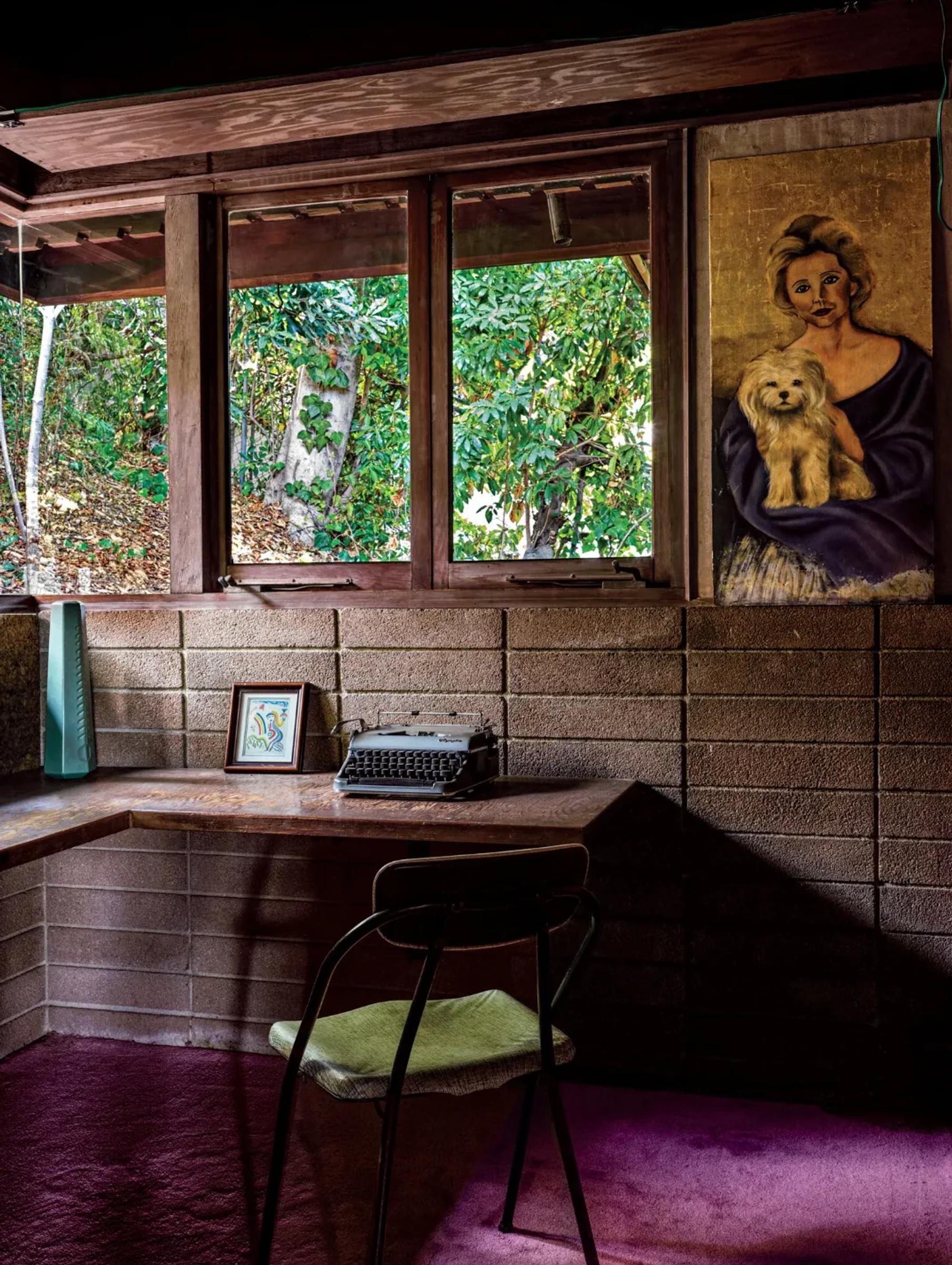A cozy Los Angeles home workspace with desk and brick walls. Features a vintage typewriter, a framed picture, chair, and hanging portrait of a woman and dog. Greenery is visible through the window.