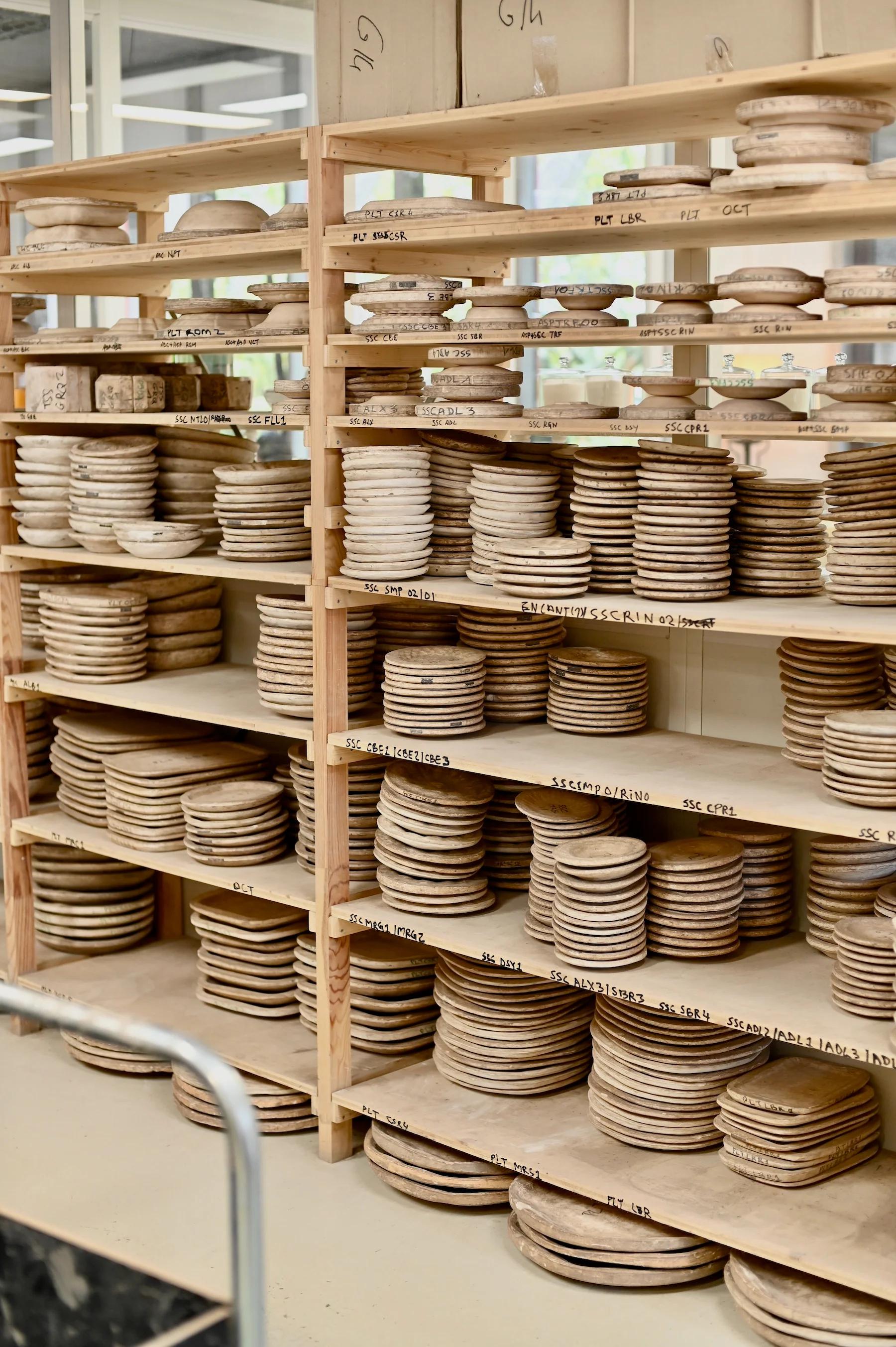 Shelves filled with stacked circular clay plates at varying drying stages. Handwritten labels indicate different processes or stages.