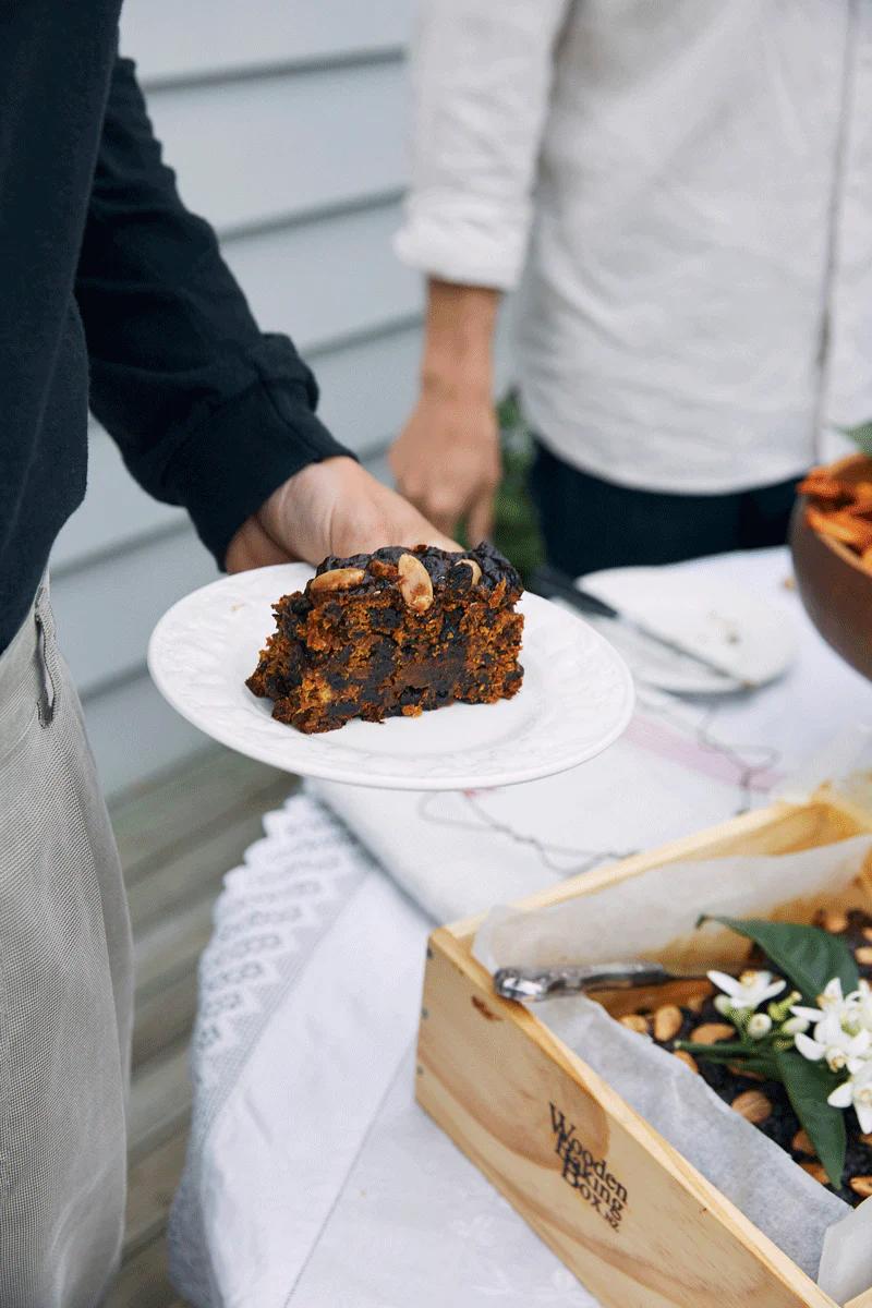 Person holding plate with fruit cake slice. Table with lace tablecloth and various foods in background.