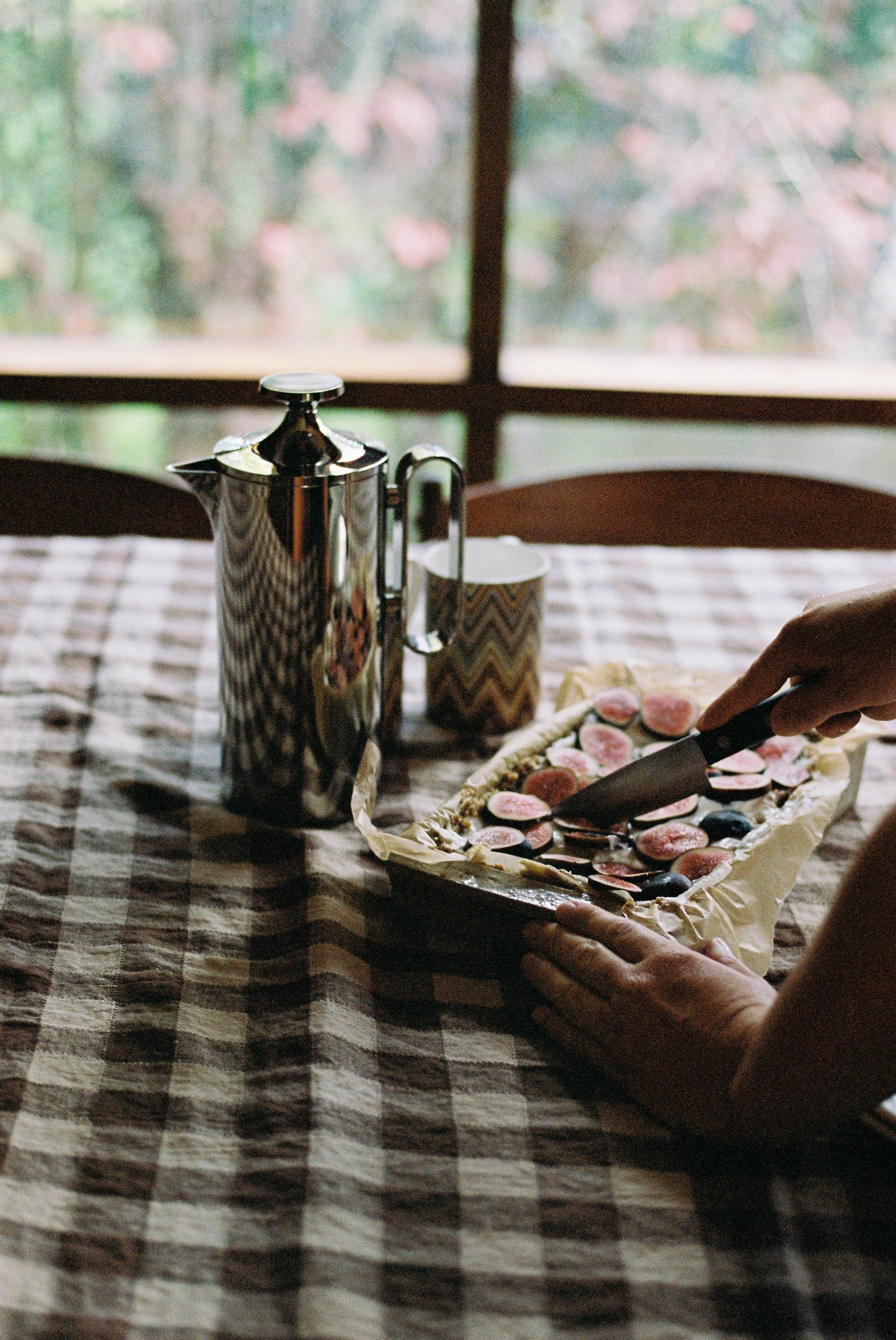 Person spreading fig slices on parchment-lined tray. Coffee pot and mug nearby. Checkered tablecloth.