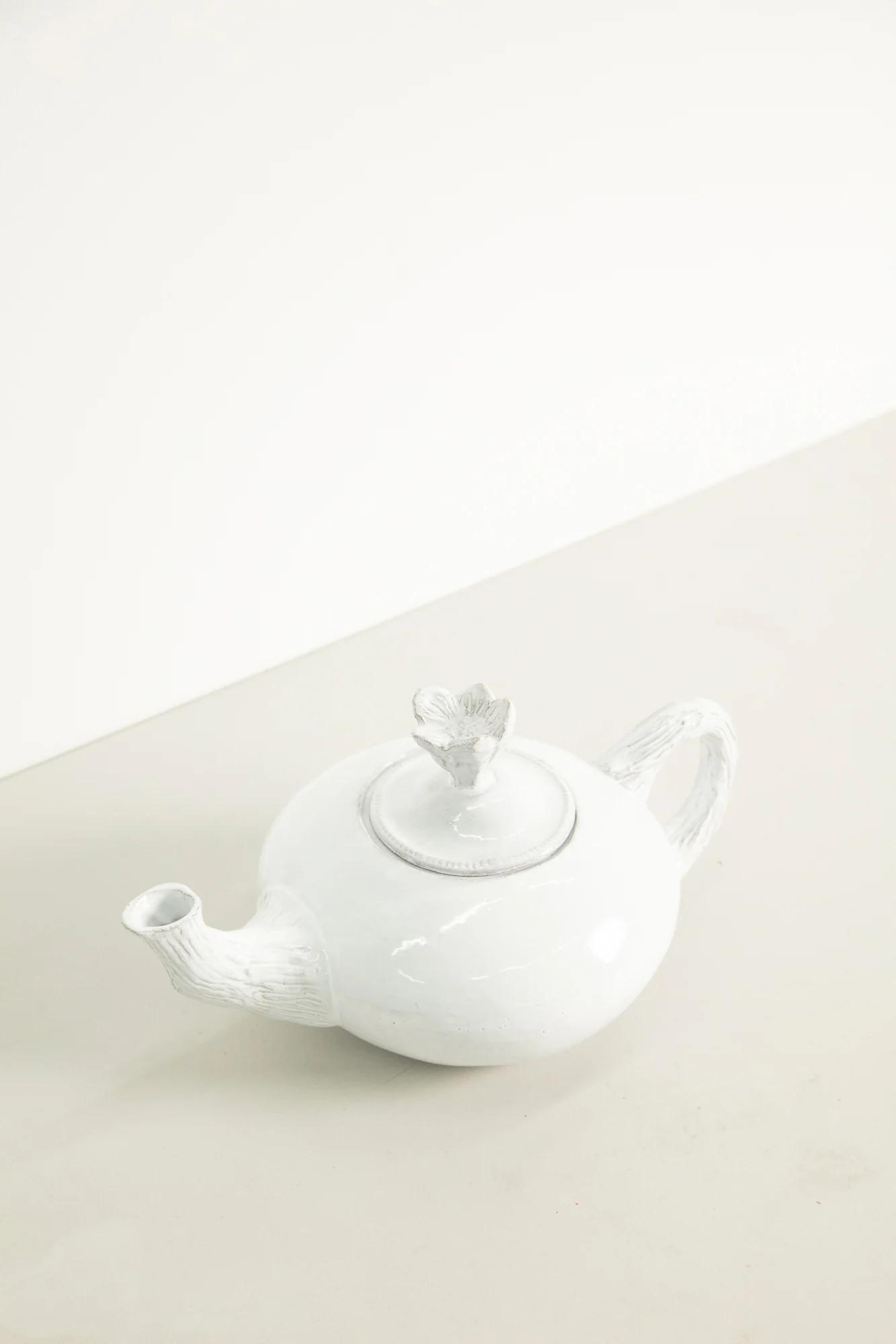 White ceramic teapot with decorative handle, spout, and lid ornament on light surface. Plain, light wall background.