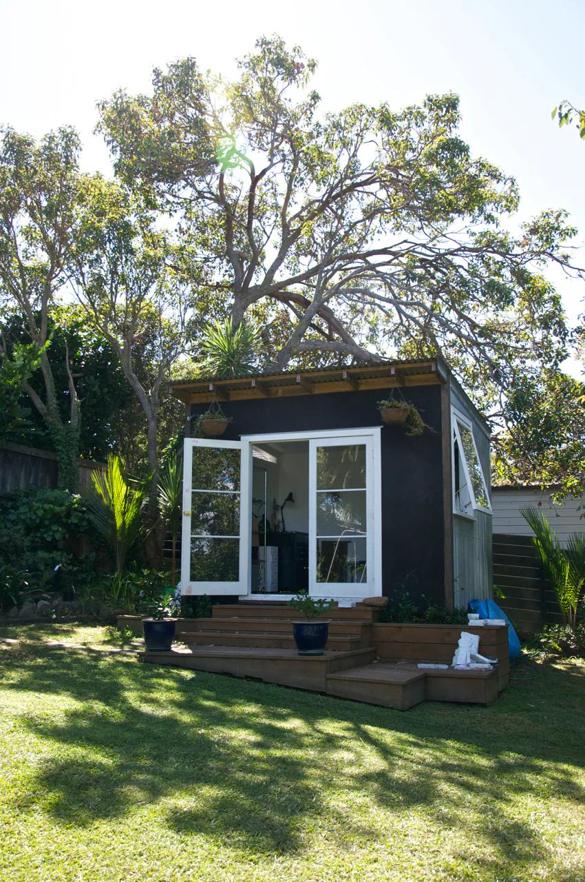 Modern shed with large windows and deck in sunny backyard. Surrounded by lush greenery, steps lead to entrance with potted plants on deck.