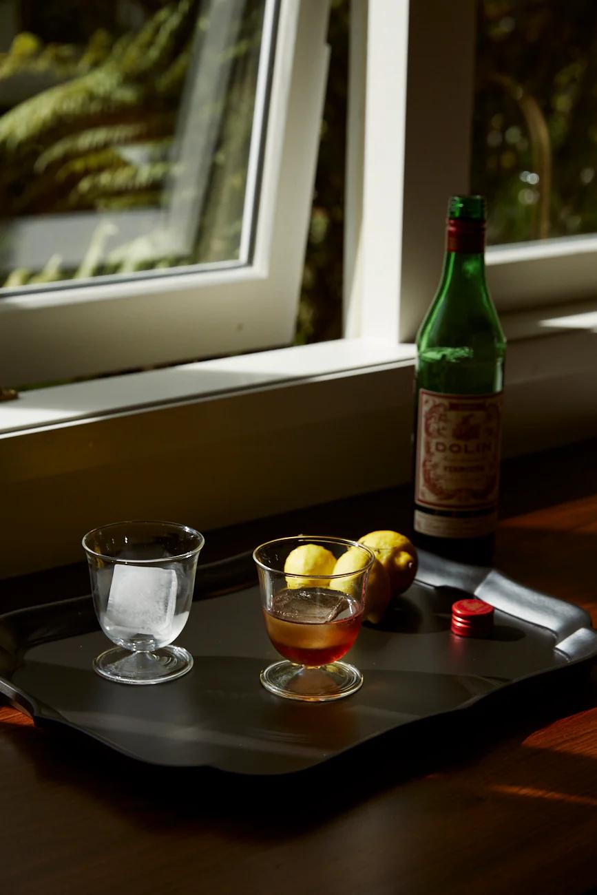 A sunlit tray holds a green Vermouth bottle, glasses with drinks, and lemons. An open window reveals greenery, blending classic and modern Italian design.