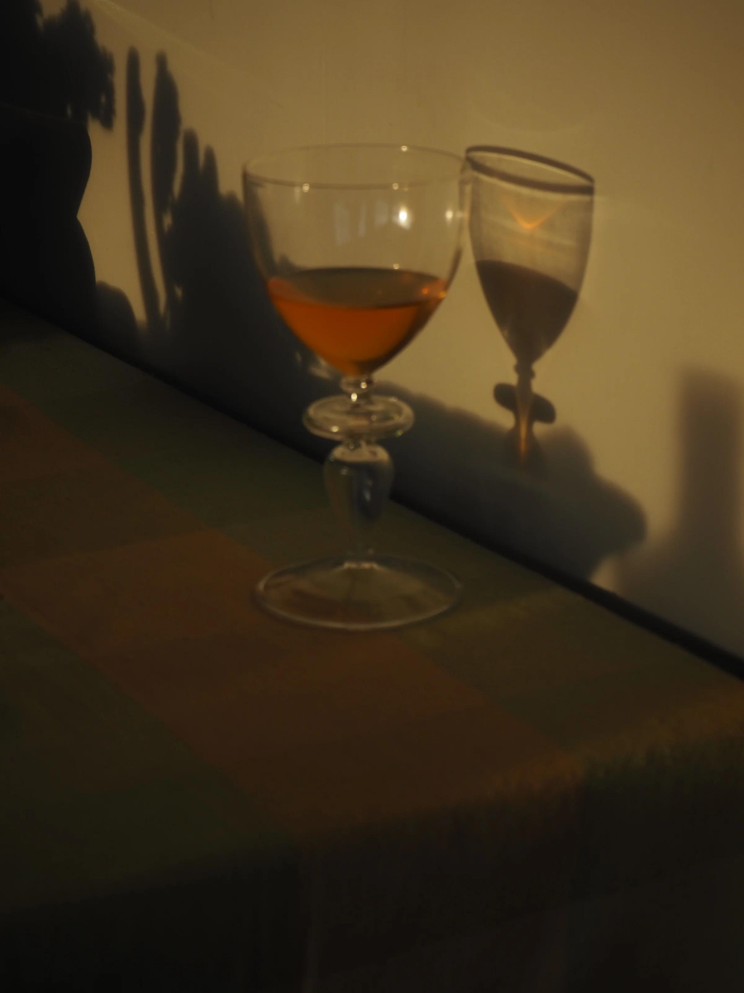 Amber natural wine in glass on table casts shadow on wall. Light from right creates soft, warm tones in subdued setting, emphasising glass details.