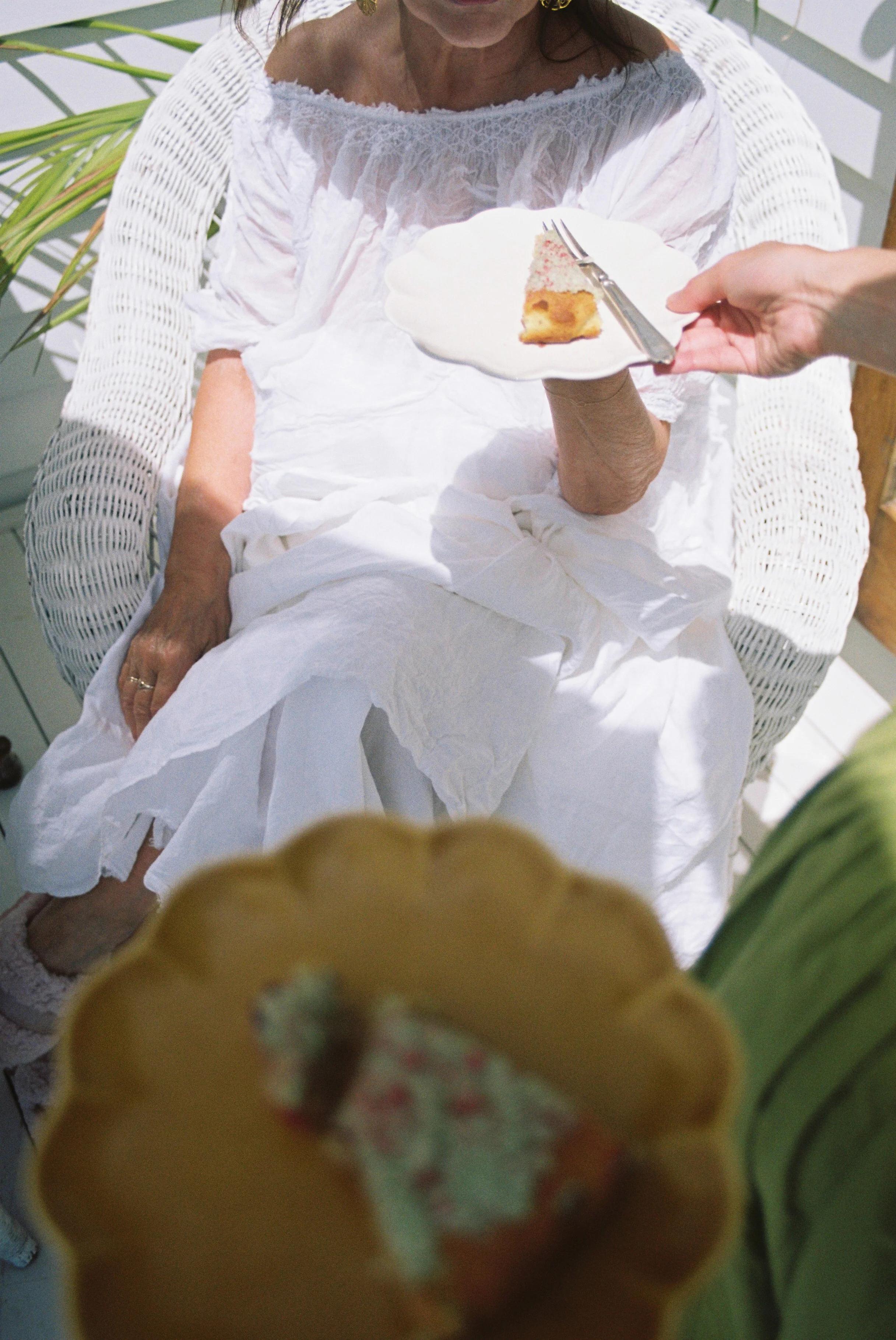 Woman in white dress on wicker chair holding pizza slice. Hand offering utensils. Blurred plate in foreground.